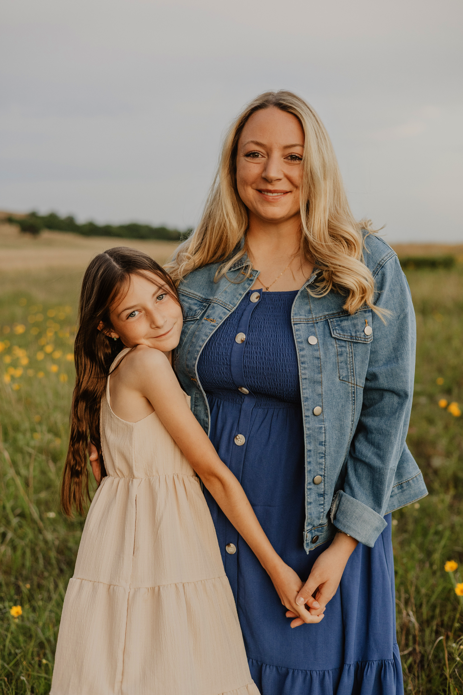 A woman in a blue dress and denim jacket poses with a young girl in a field of wildflowers at golden hour.