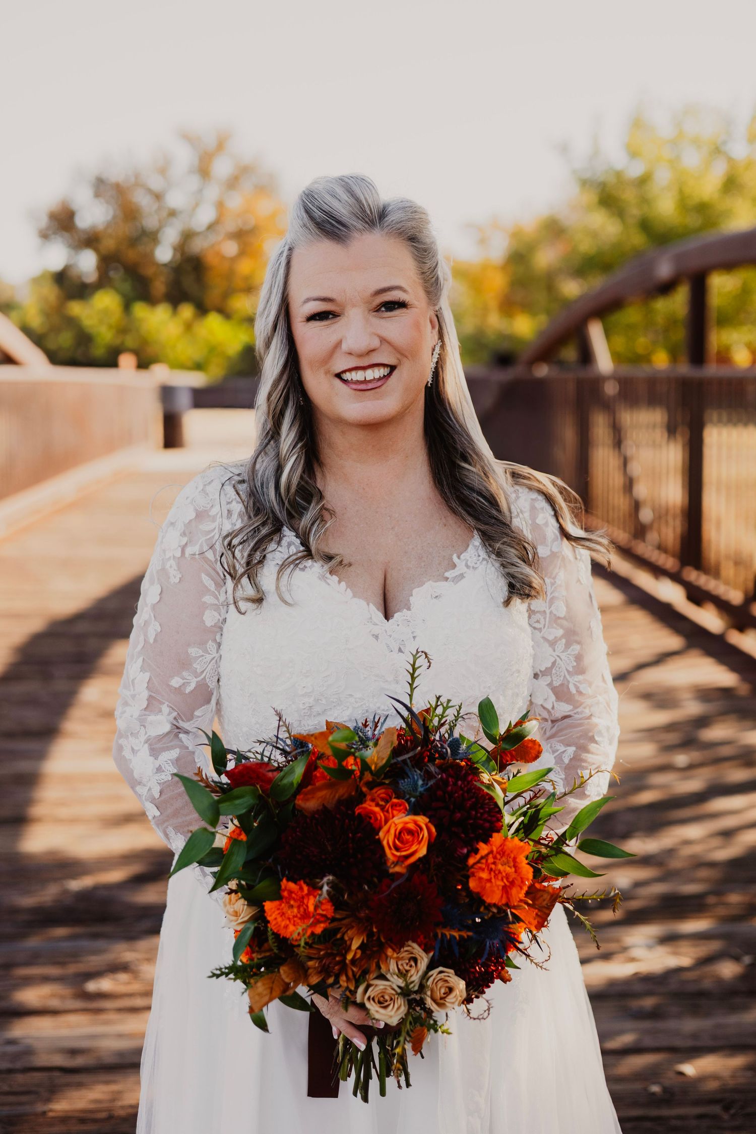 A bride in a lace wedding dress holds a vibrant fall-colored bouquet on a wooden bridge.