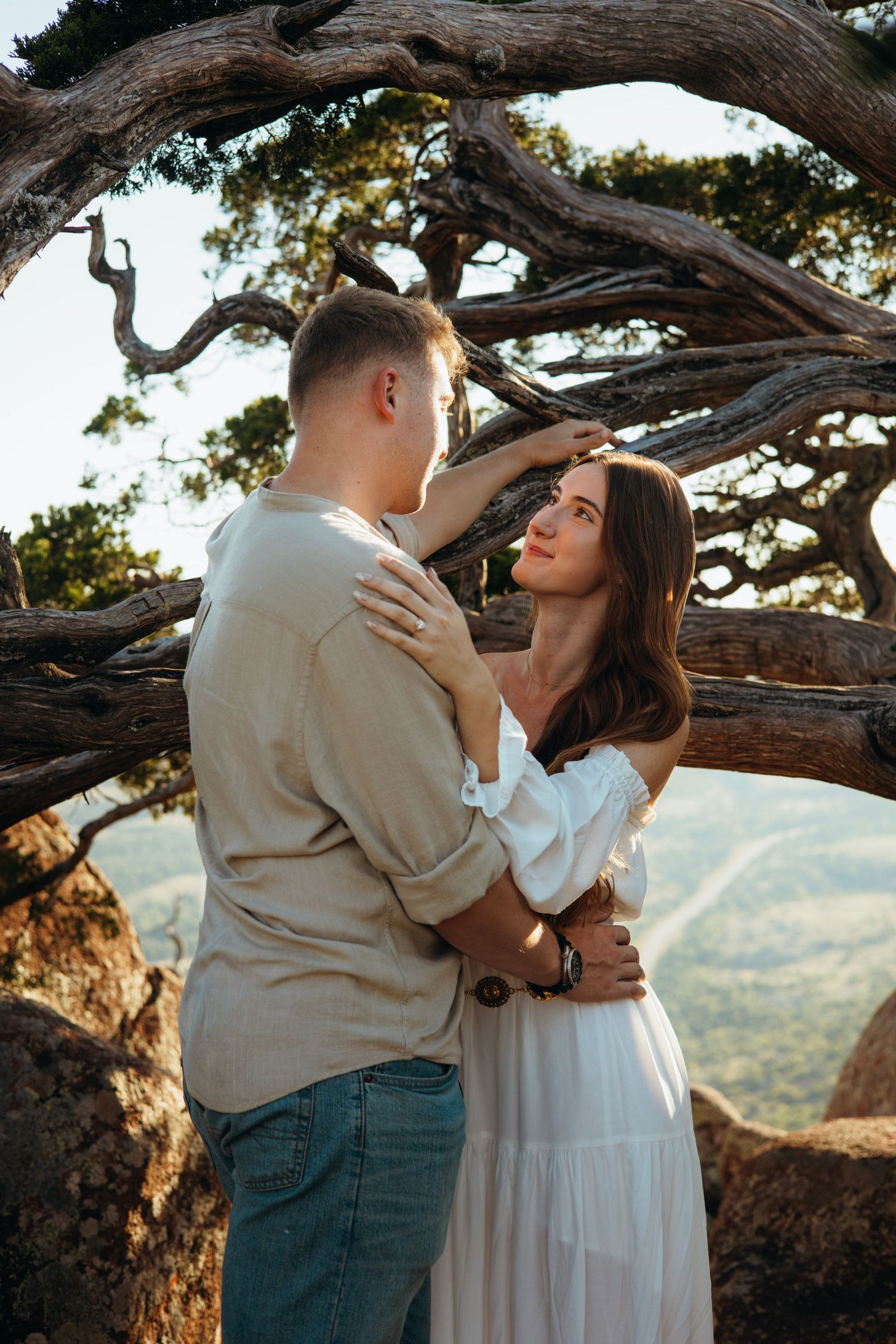A romantic couple embracing under a twisted tree branch with mountains in the background.