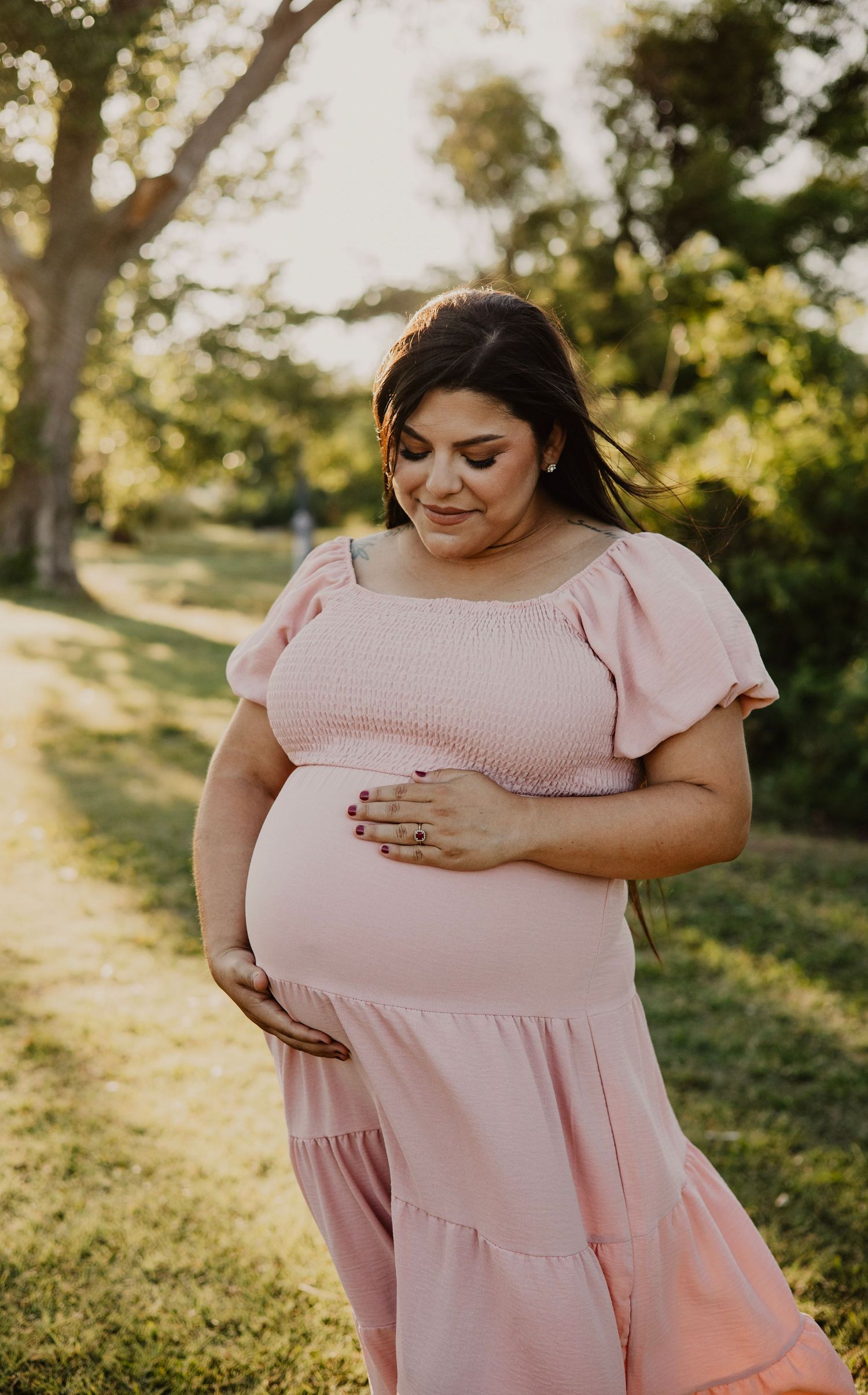 An expectant mother in a pink dress poses in a sunlit park setting.