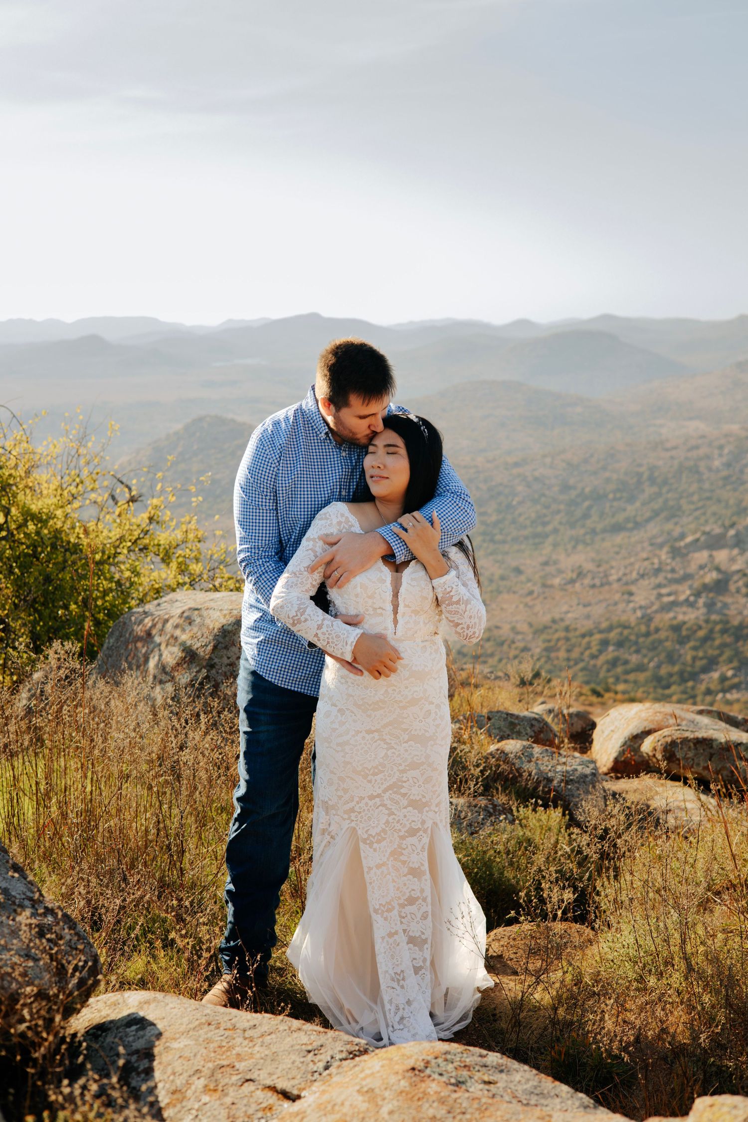 A couple embraces on rocky terrain overlooking a vast mountain landscape.