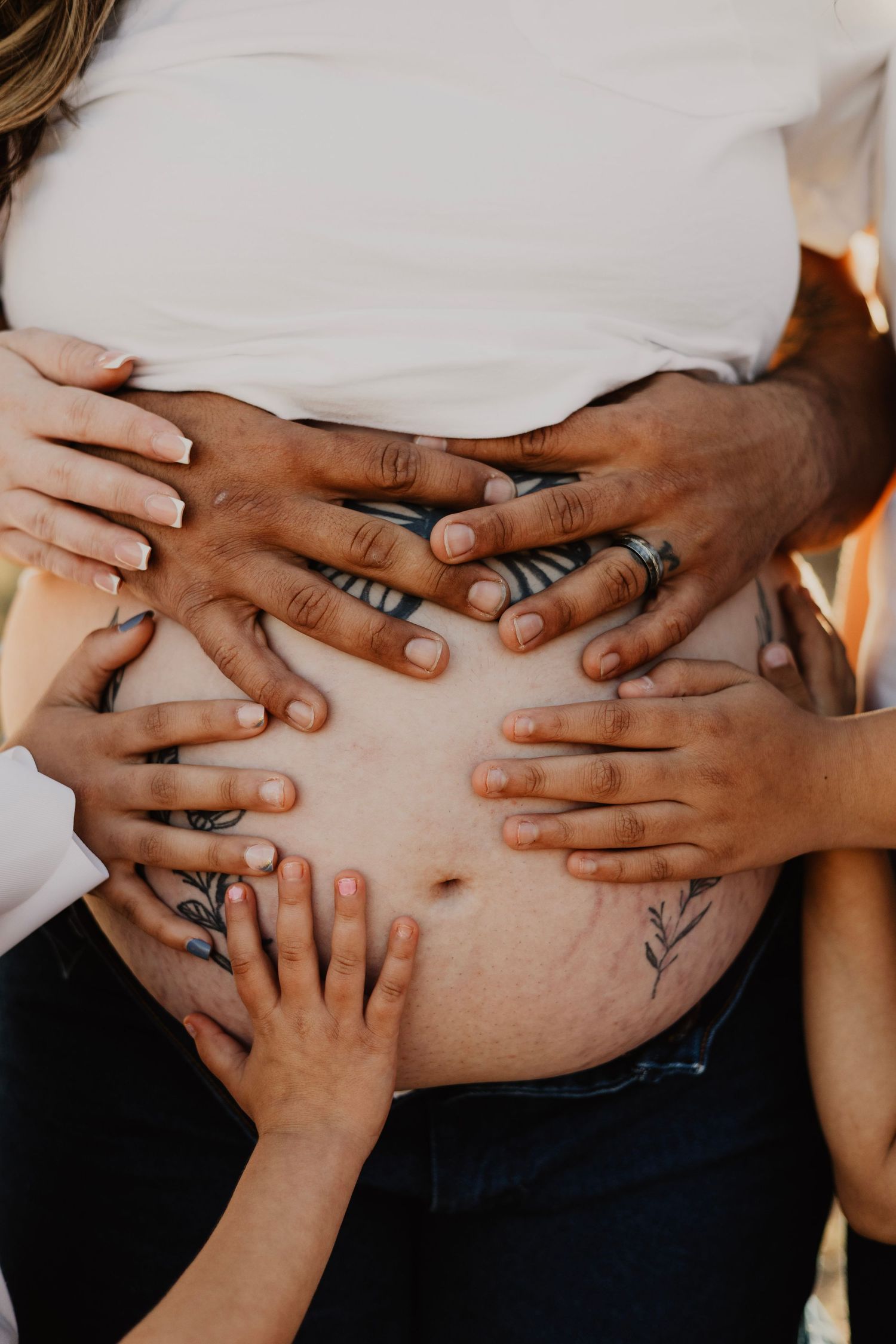 Multiple hands of a family resting on a pregnant woman's tattooed baby bump in a maternity photo.