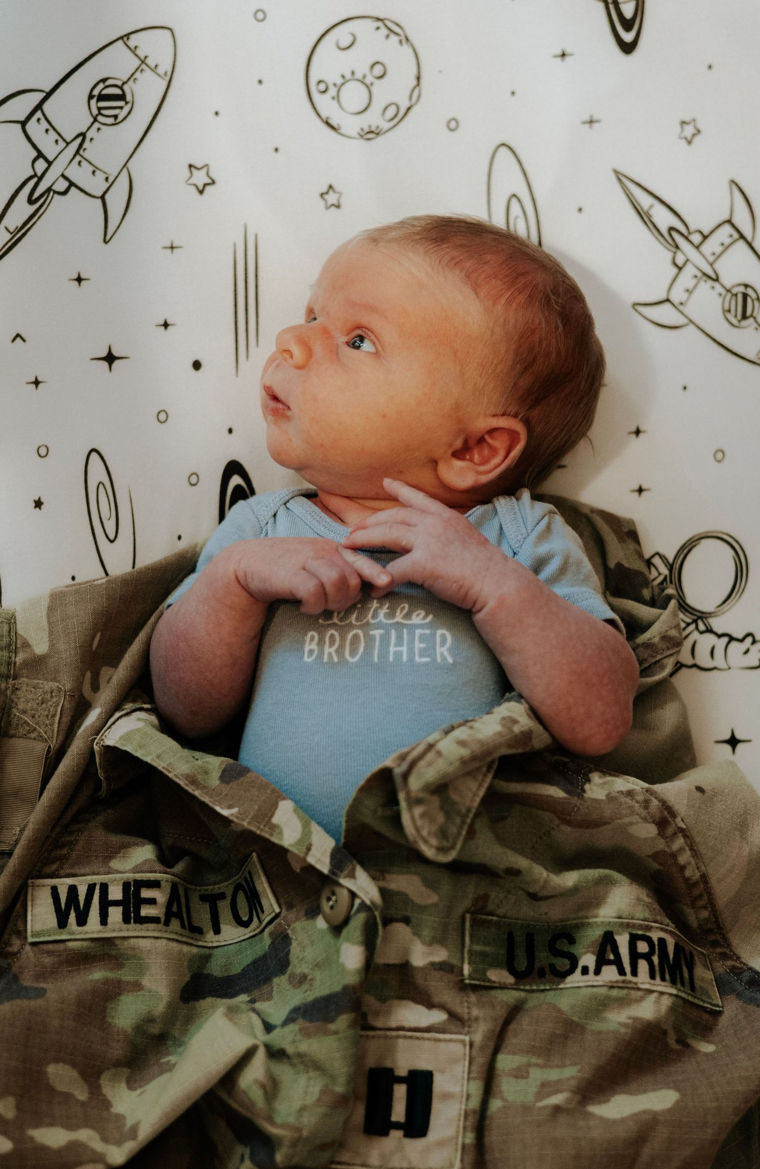 Infant in blue onesie resting on military camouflage uniform with space-themed wall art.