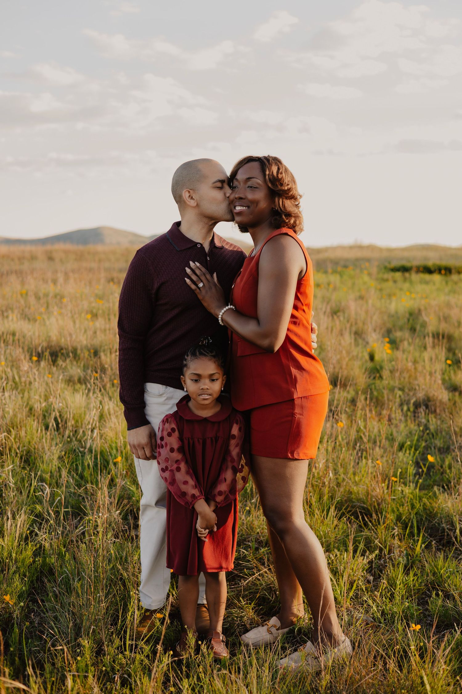 A family of three poses in a golden field at sunset, with parents sharing a tender kiss while their young daughter stands in front.