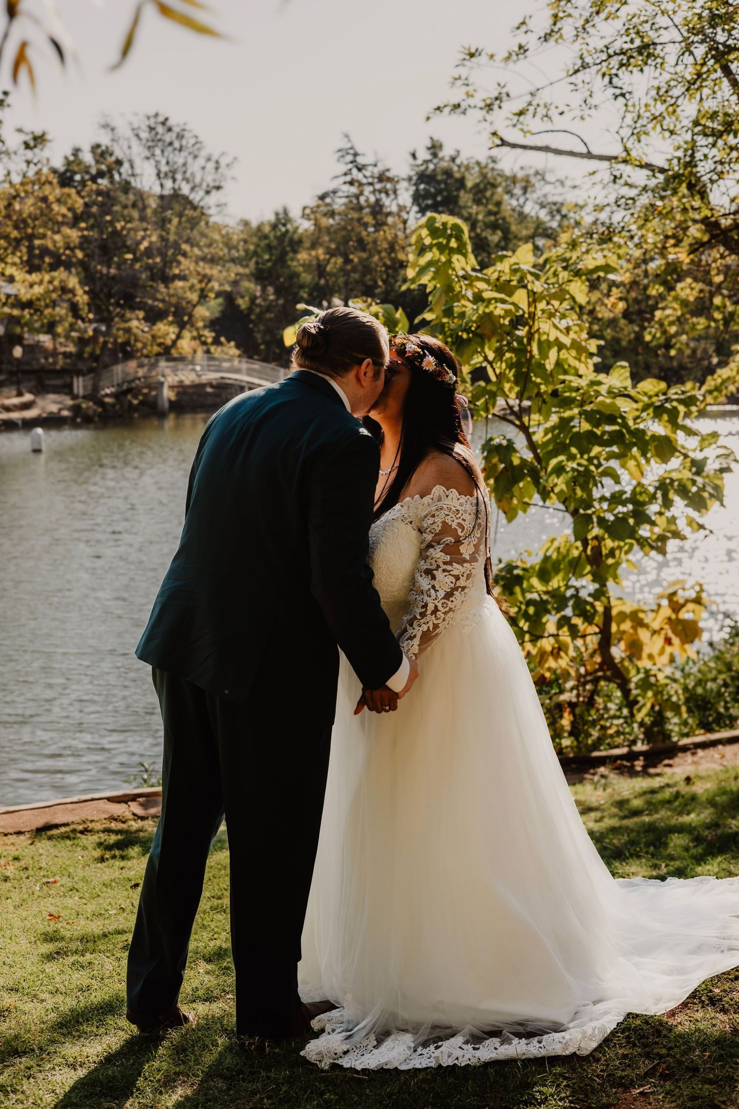 Wedding couple embraces by lakeside with autumn trees in background.