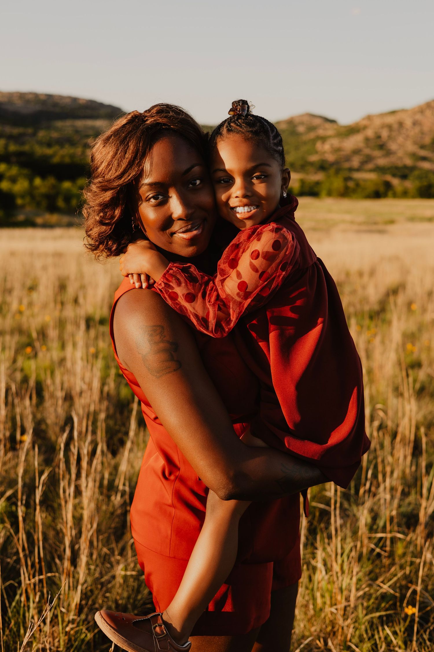 A mother in a red dress holds her smiling daughter in a sunlit field with hills in the background.