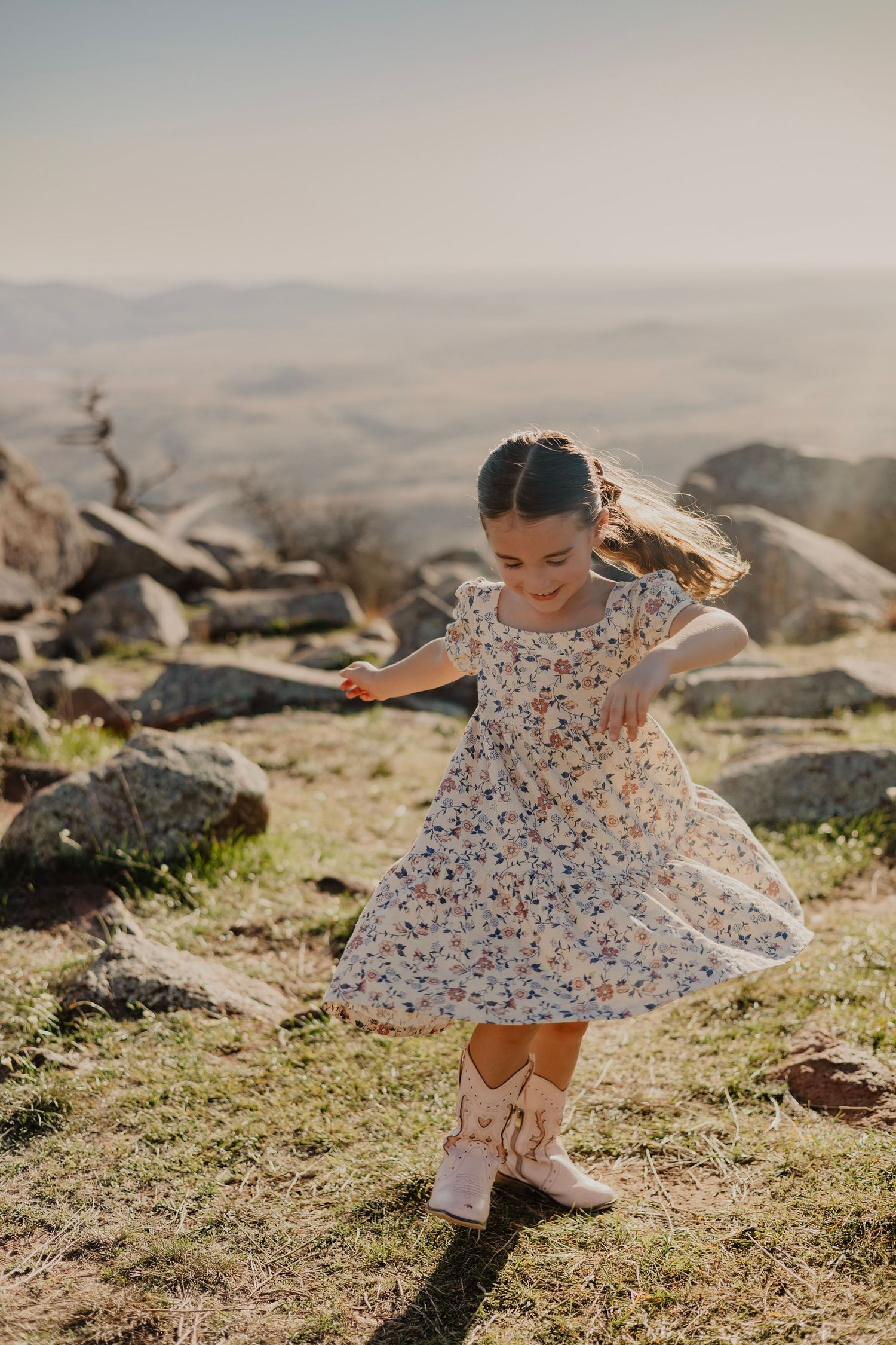 Young girl twirling in floral dress and cowboy boots on rocky hilltop with scenic valley views.