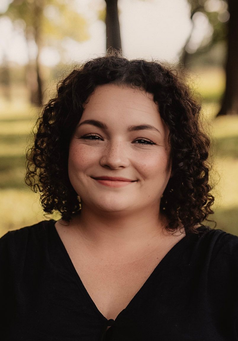 Portrait of a person with curly dark hair and a warm smile wearing a black v-neck top against a blurred outdoor background.