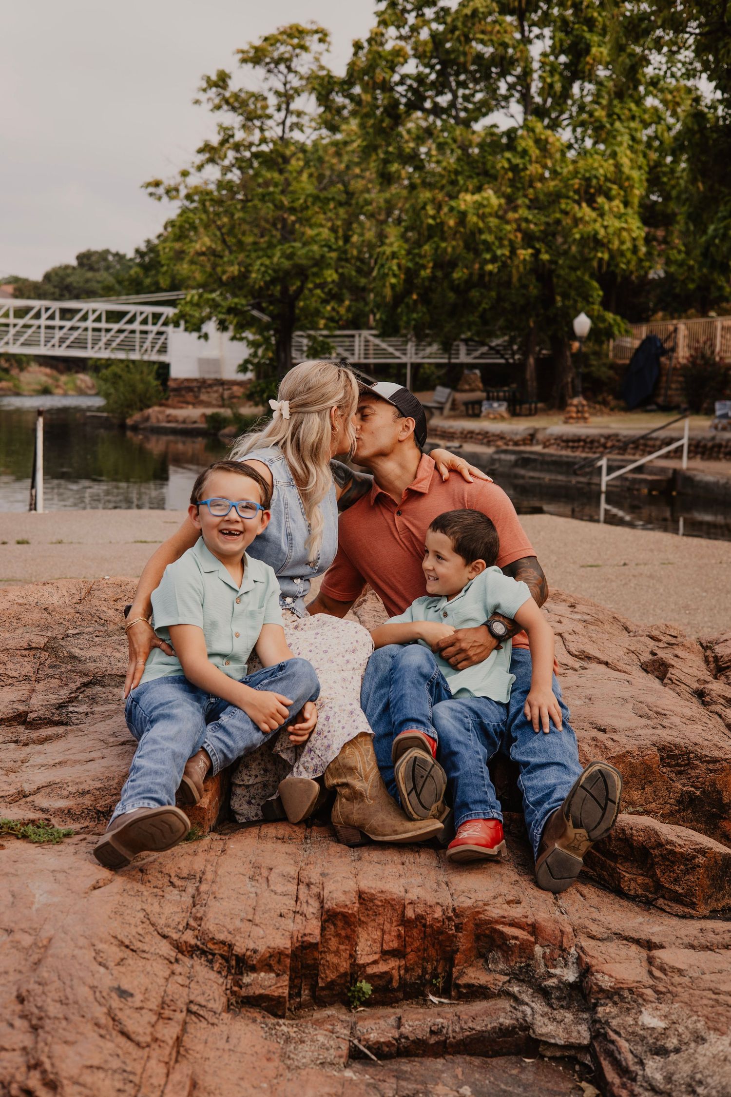 A family of four sits together on red rocks near a lake, sharing an intimate moment outdoors.