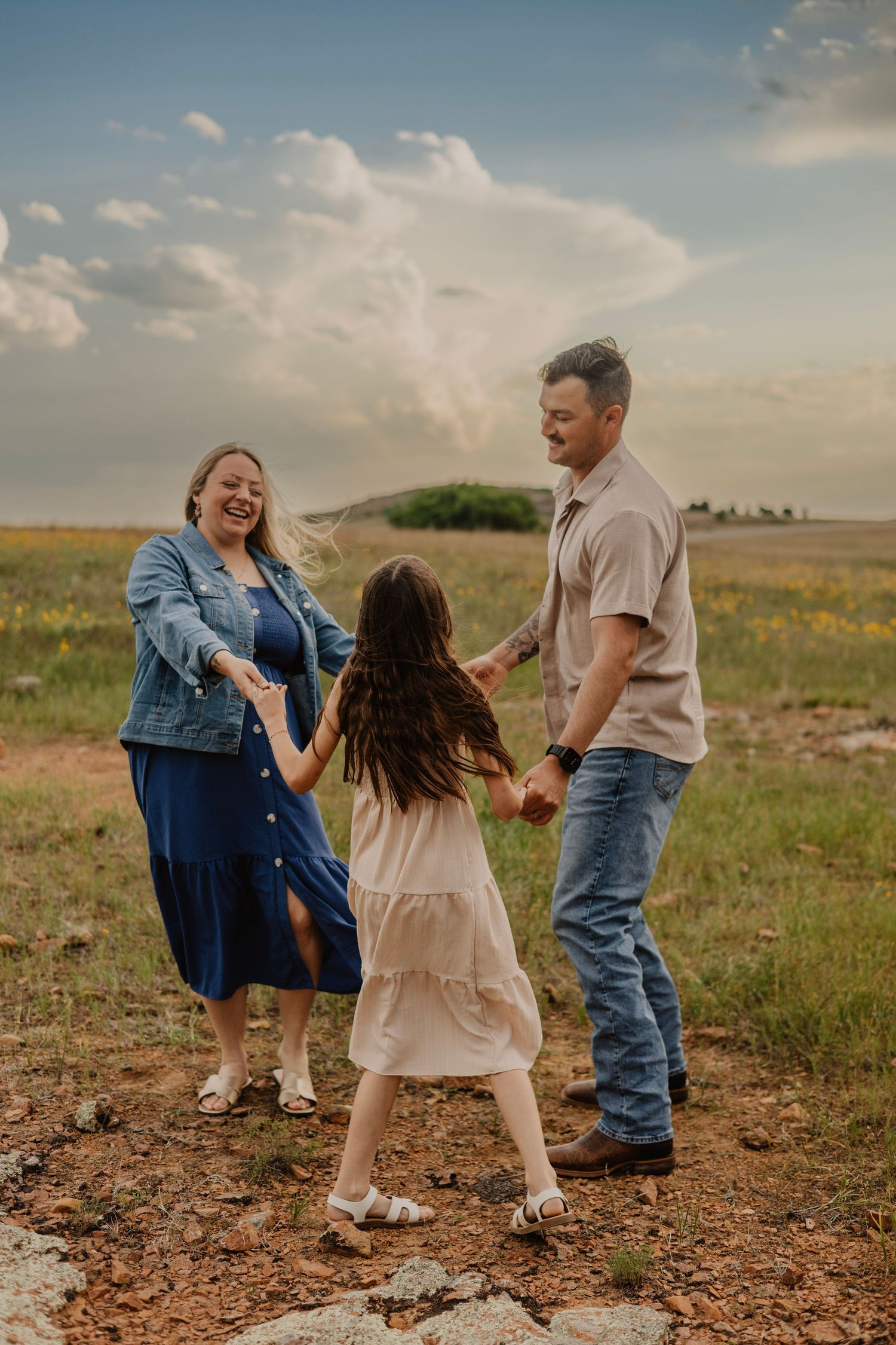 A joyful family of three holds hands and dances together in a green meadow under a dramatic cloudy sky.