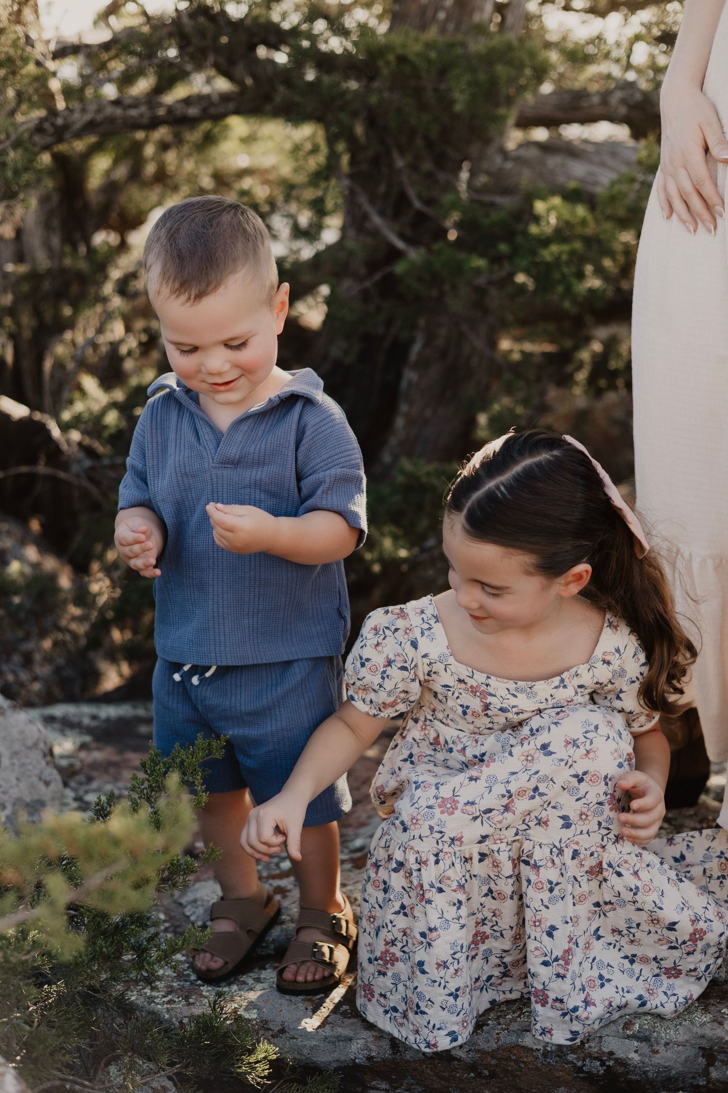 A young boy in blue outfit and a girl in floral dress play together outdoors among trees and rocks.
