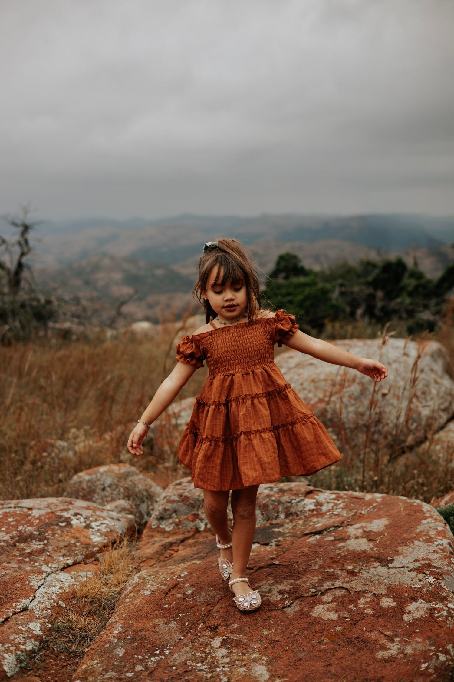 Child in rust-colored dress exploring rocky terrain against cloudy mountain backdrop.