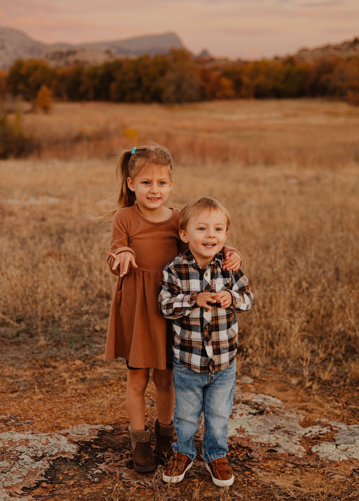 Two children in fall outfits standing together on rocks in a golden field at sunset.