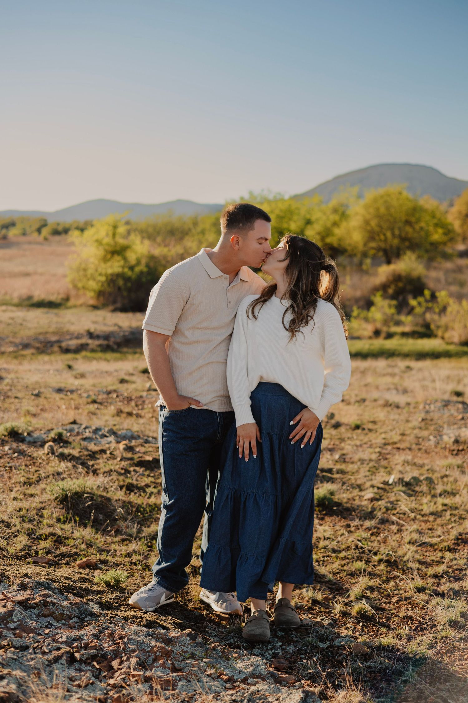 Couple sharing a kiss in a golden hour desert landscape with mountains and green shrubs in the background.