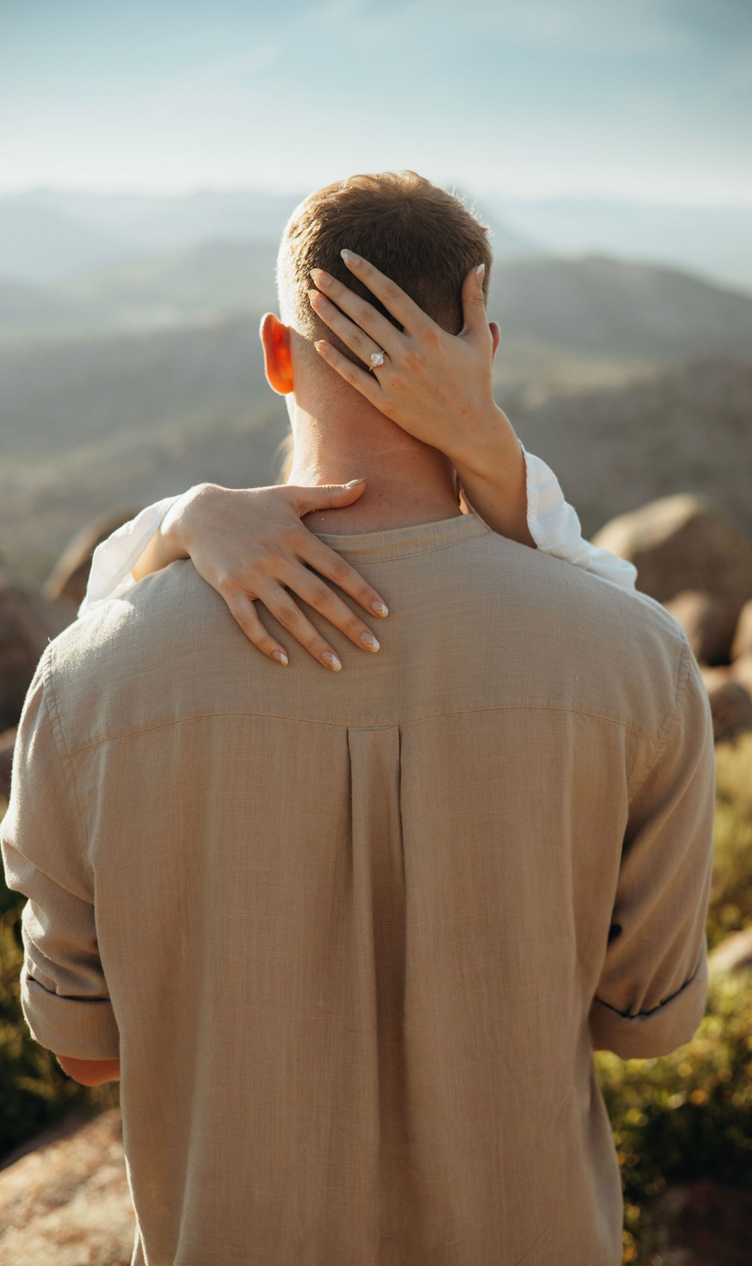 Hands tenderly embracing from behind against a mountainous backdrop.