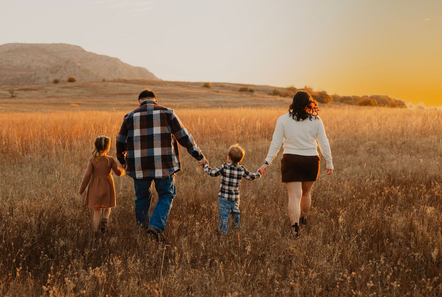 Family walking together through golden wheat field at sunset with mountains in background.