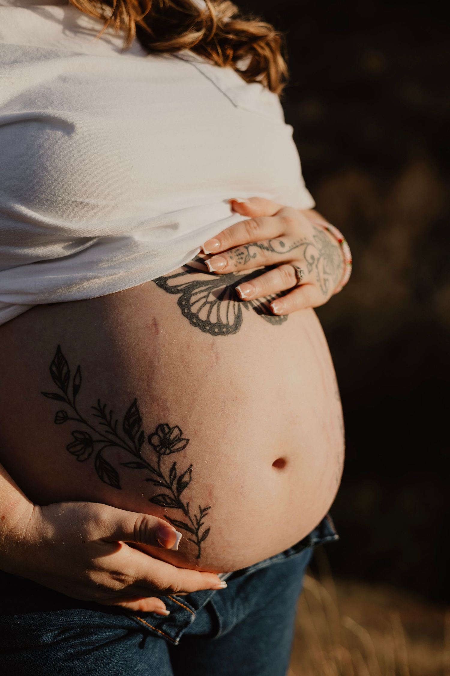 Pregnant woman with floral and butterfly tattoos cradling her bare baby bump outdoors in warm sunlight.