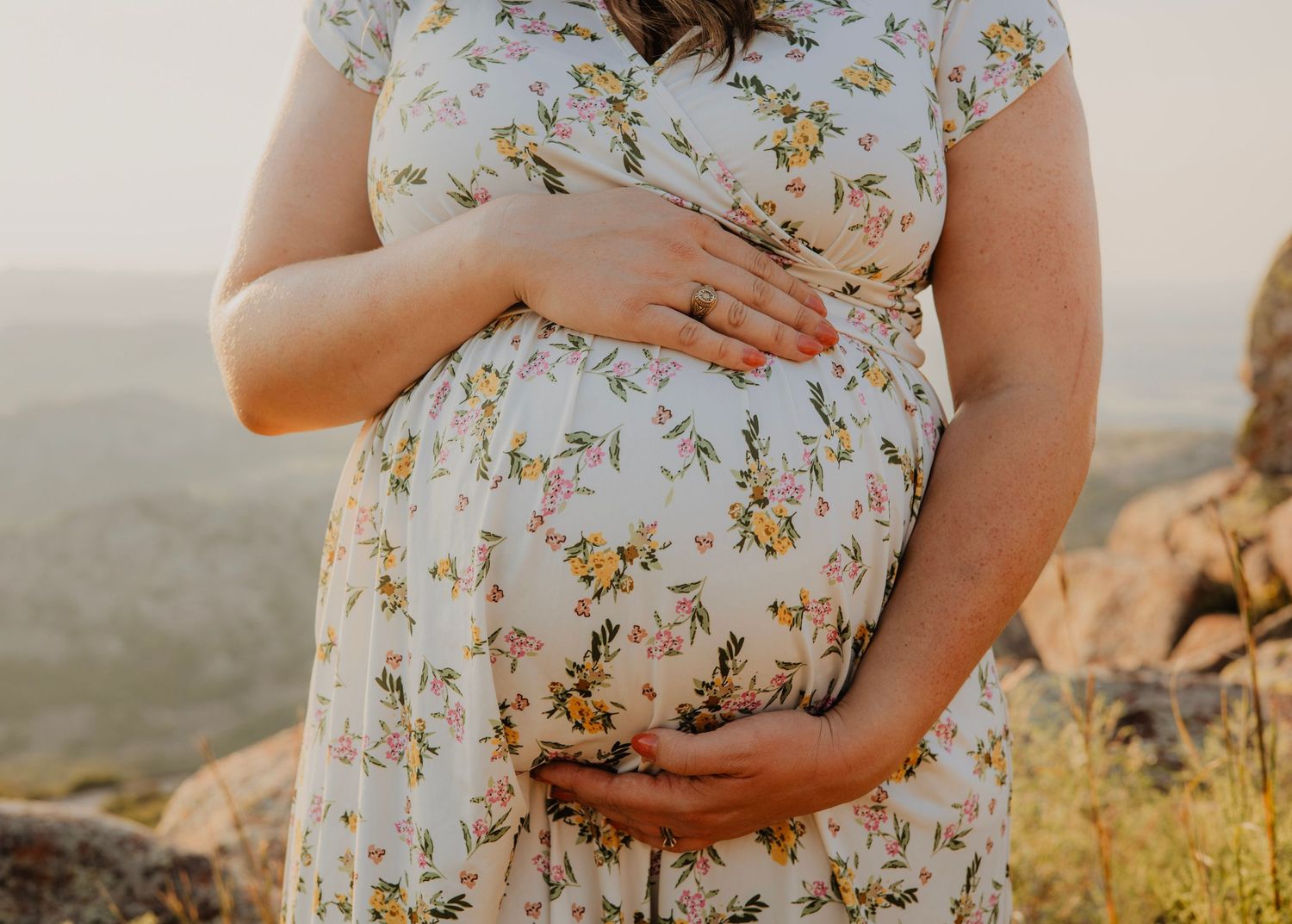 Close-up of hands cradling a pregnant belly in a floral dress against a mountain landscape.