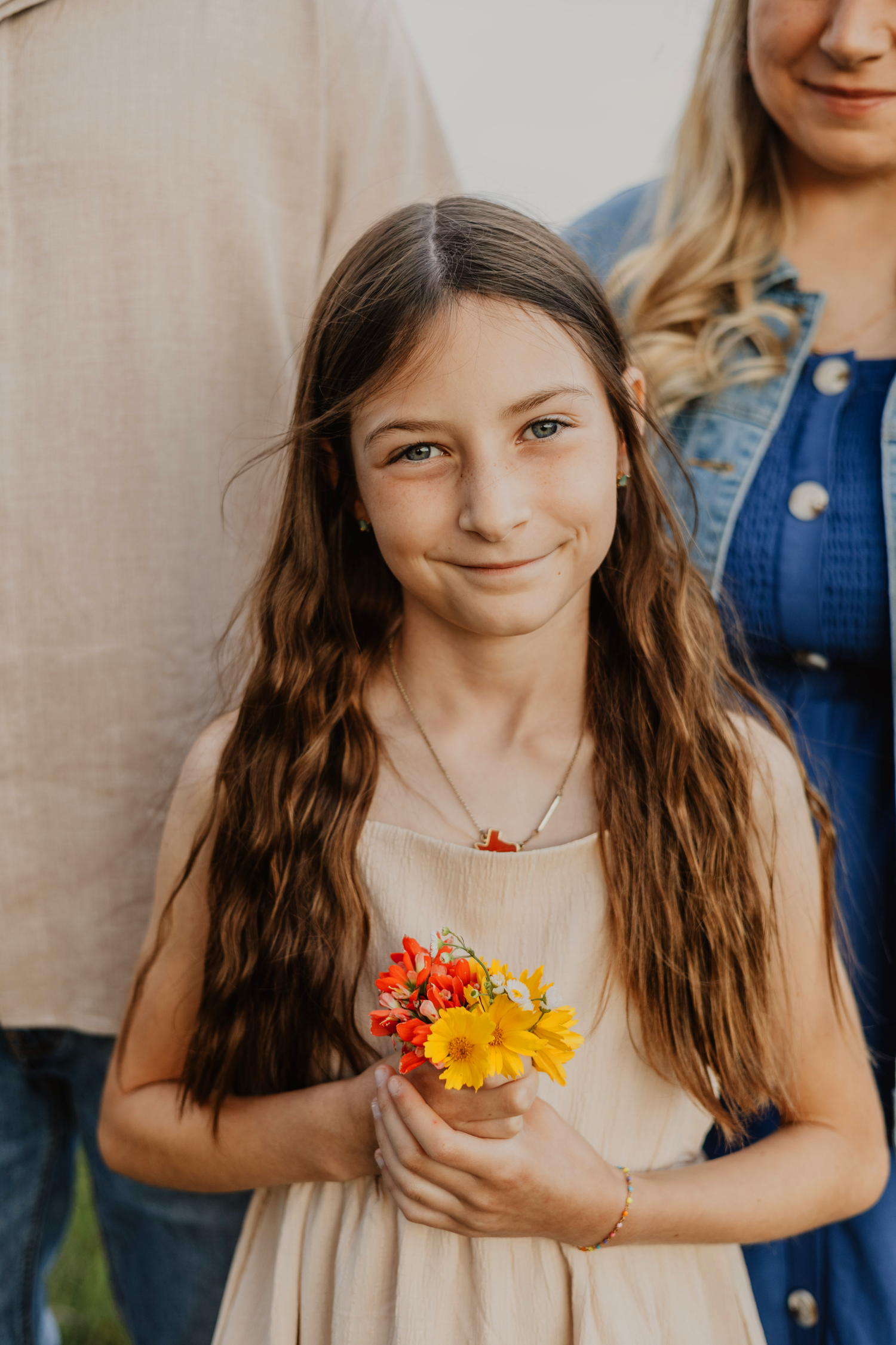 A young girl with long brown hair holds a small bouquet of colorful wildflowers, smiling softly in a beige dress.