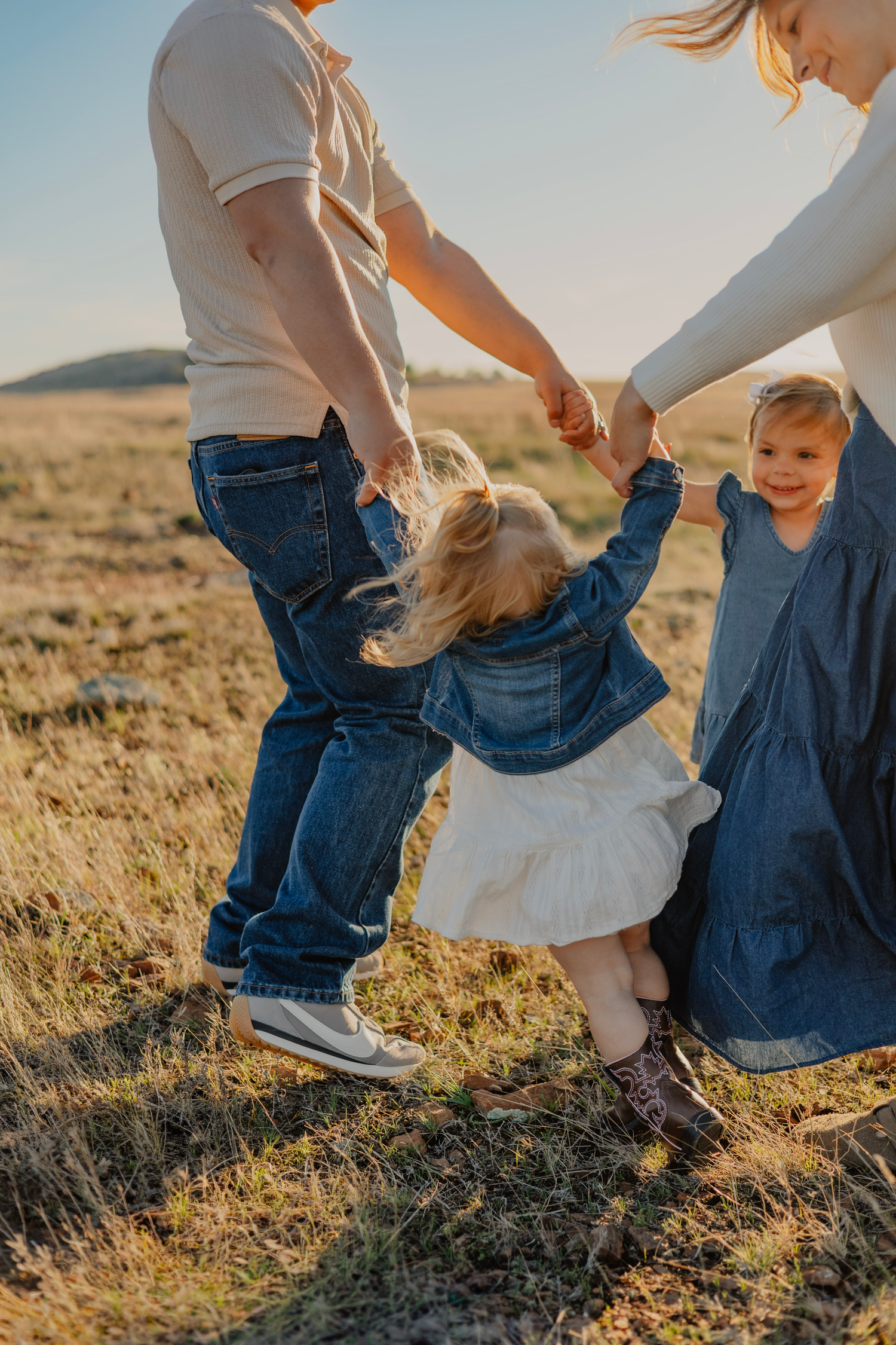 A joyful family swings their toddler daughter between them in a sunny golden field, all dressed in denim and casual wear.