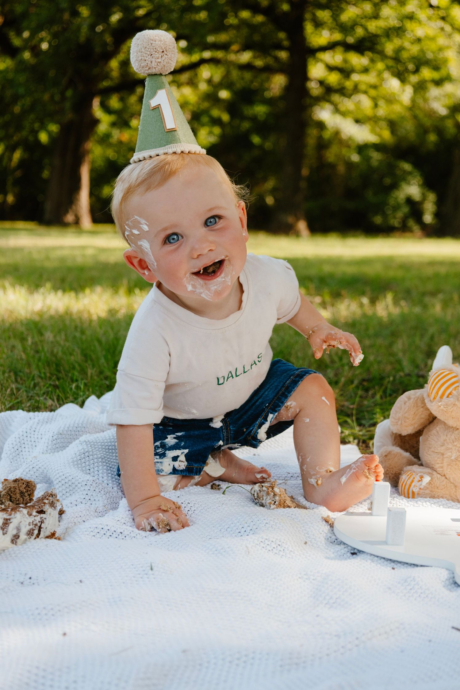 Baby wearing party hat sits on blanket with birthday cake and decorations.