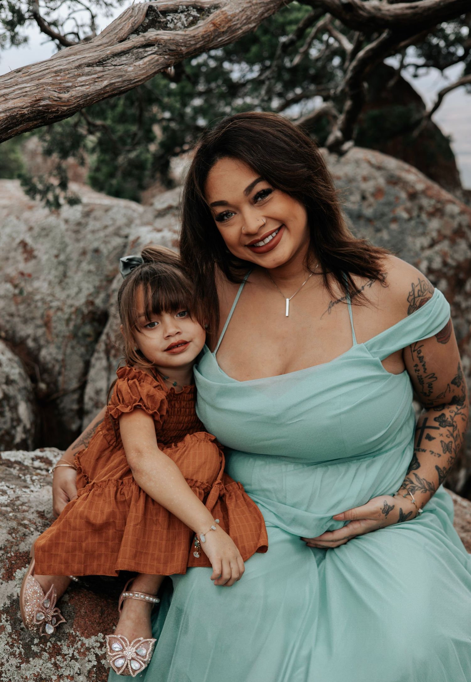 A woman in mint dress and child in orange dress pose together on rocky mountain terrain.