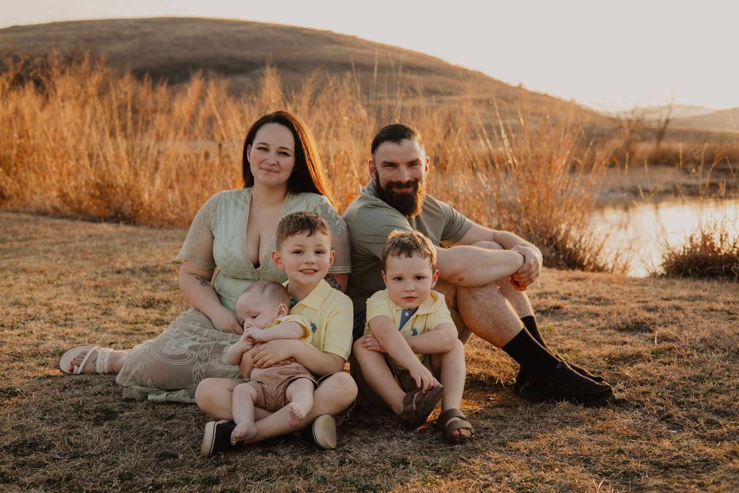 Family sits together on grassy hill during golden hour sunset near a lake.