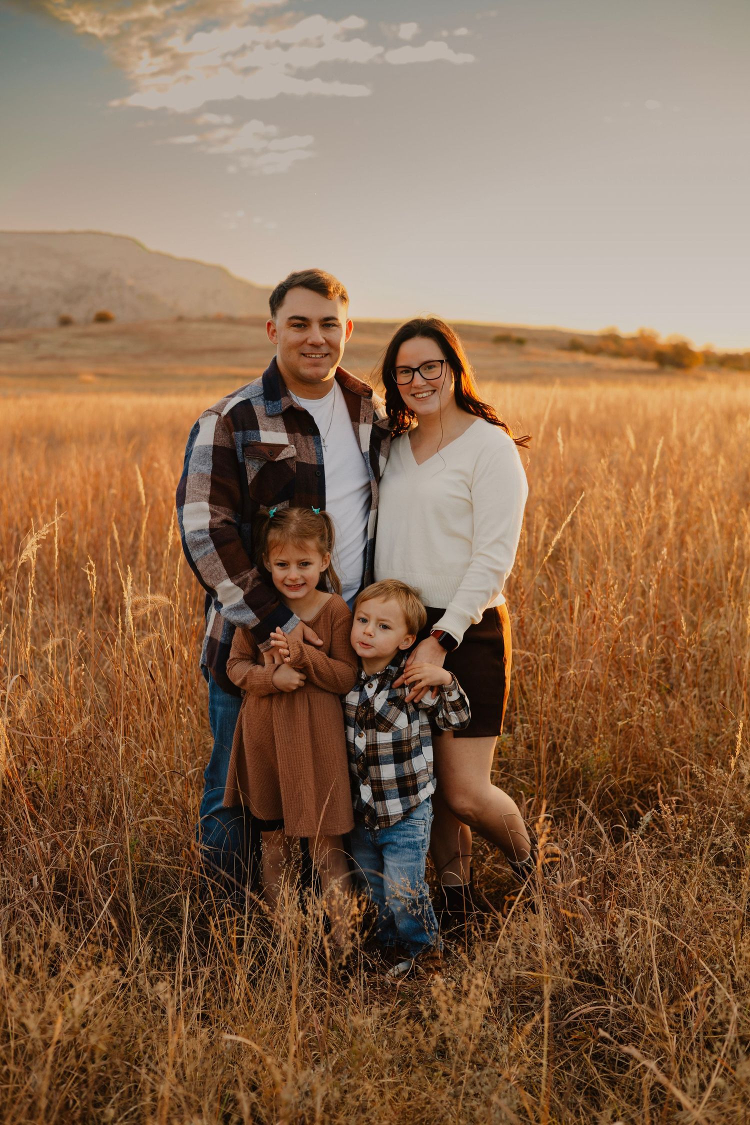 Family of four posing in golden field during sunset with mountains in background.