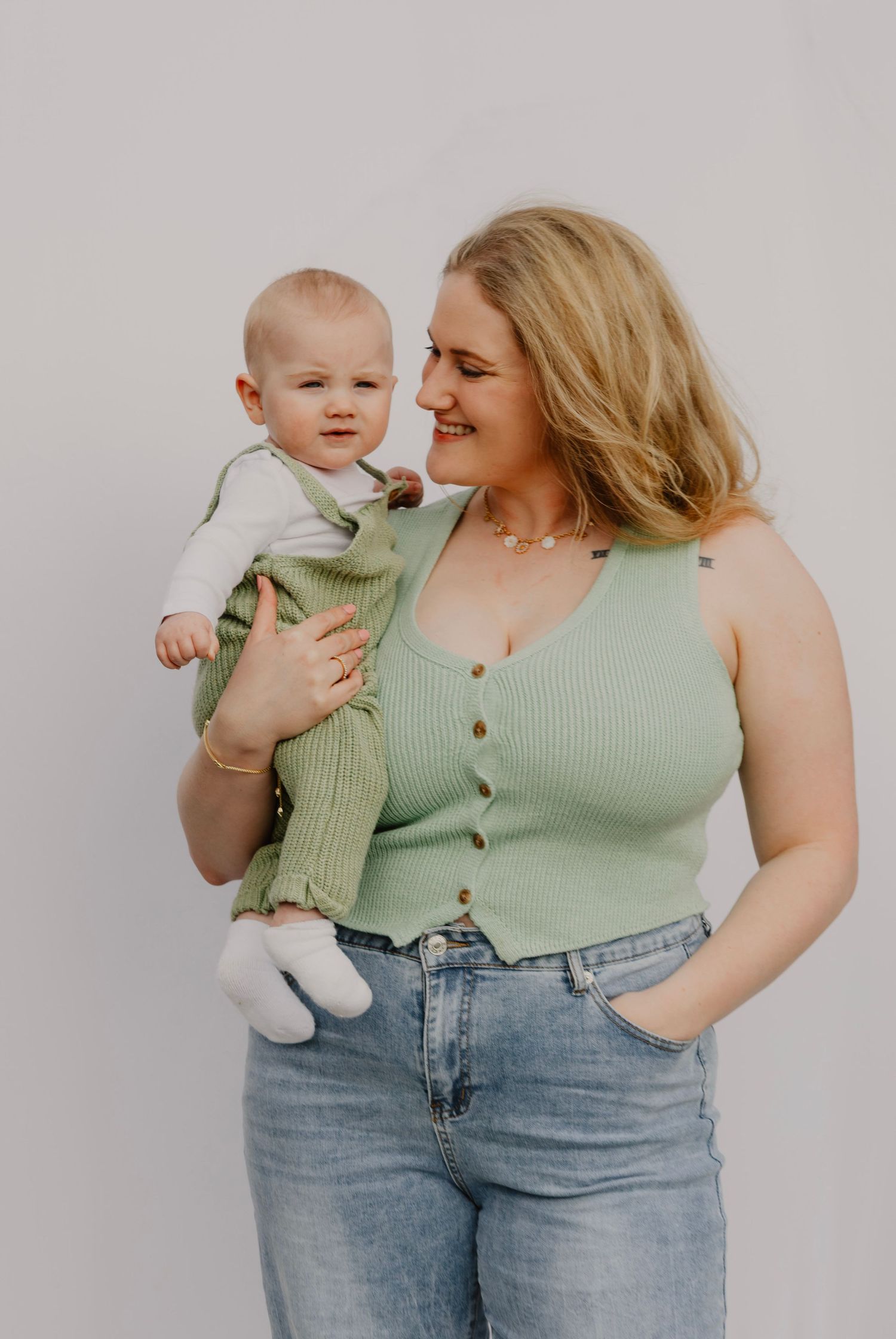 Parent in mint green top holds baby wearing gray outfit against white background.