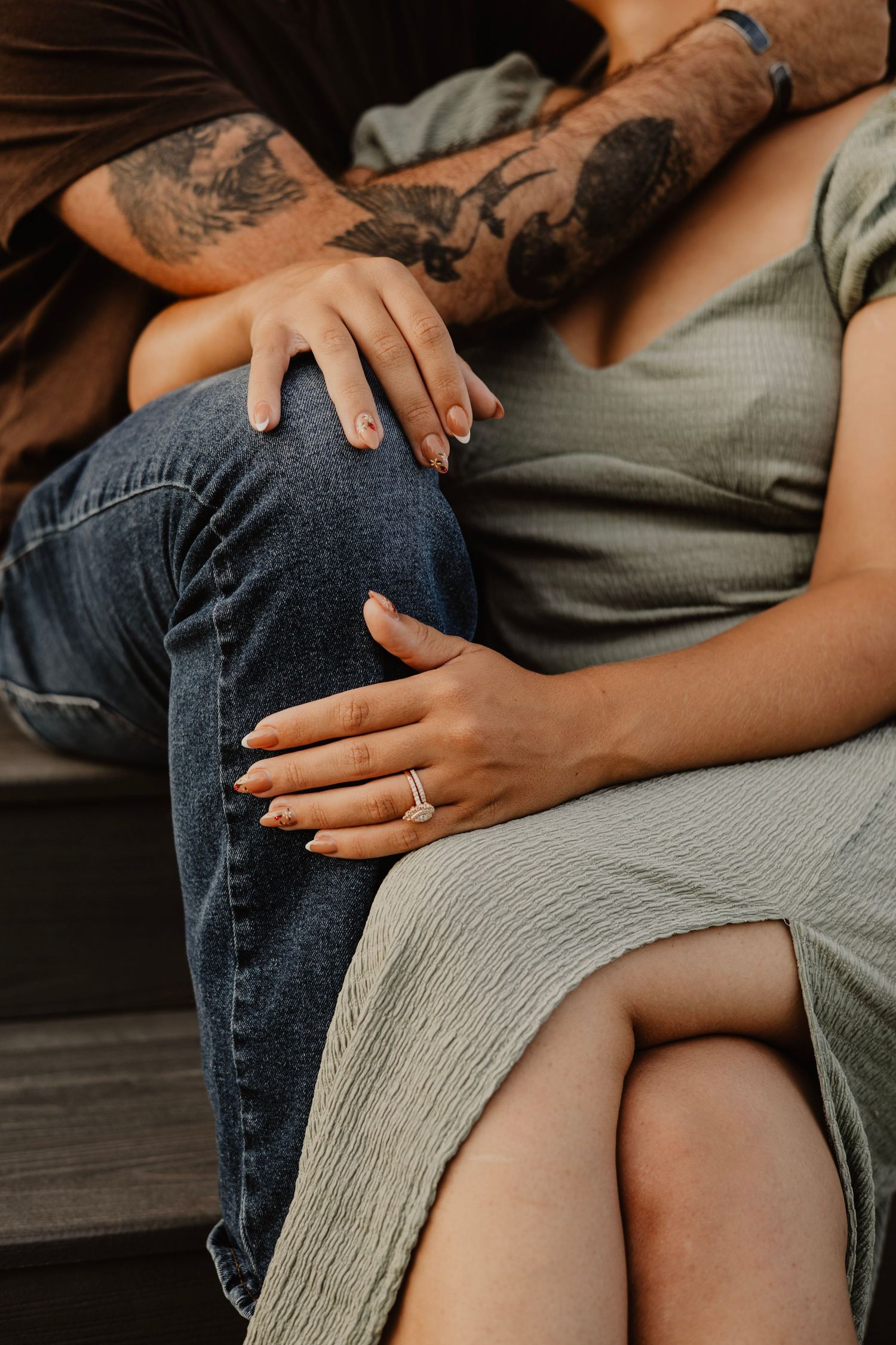 Close-up of tattooed arm and hands resting on denim jeans and sage green dress.