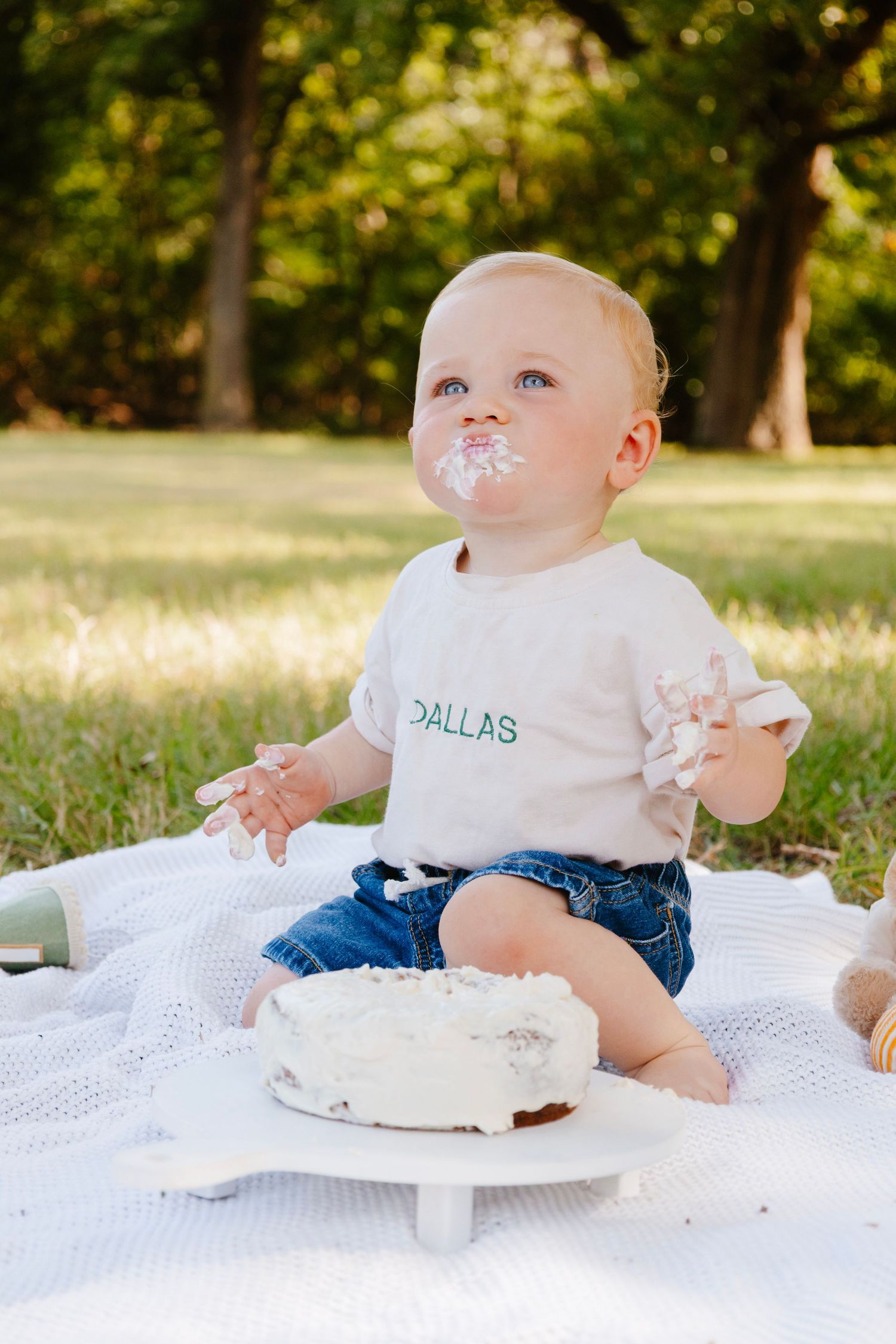 Baby in Dallas shirt with cake smeared on face sits on white blanket during cake smash photo session.