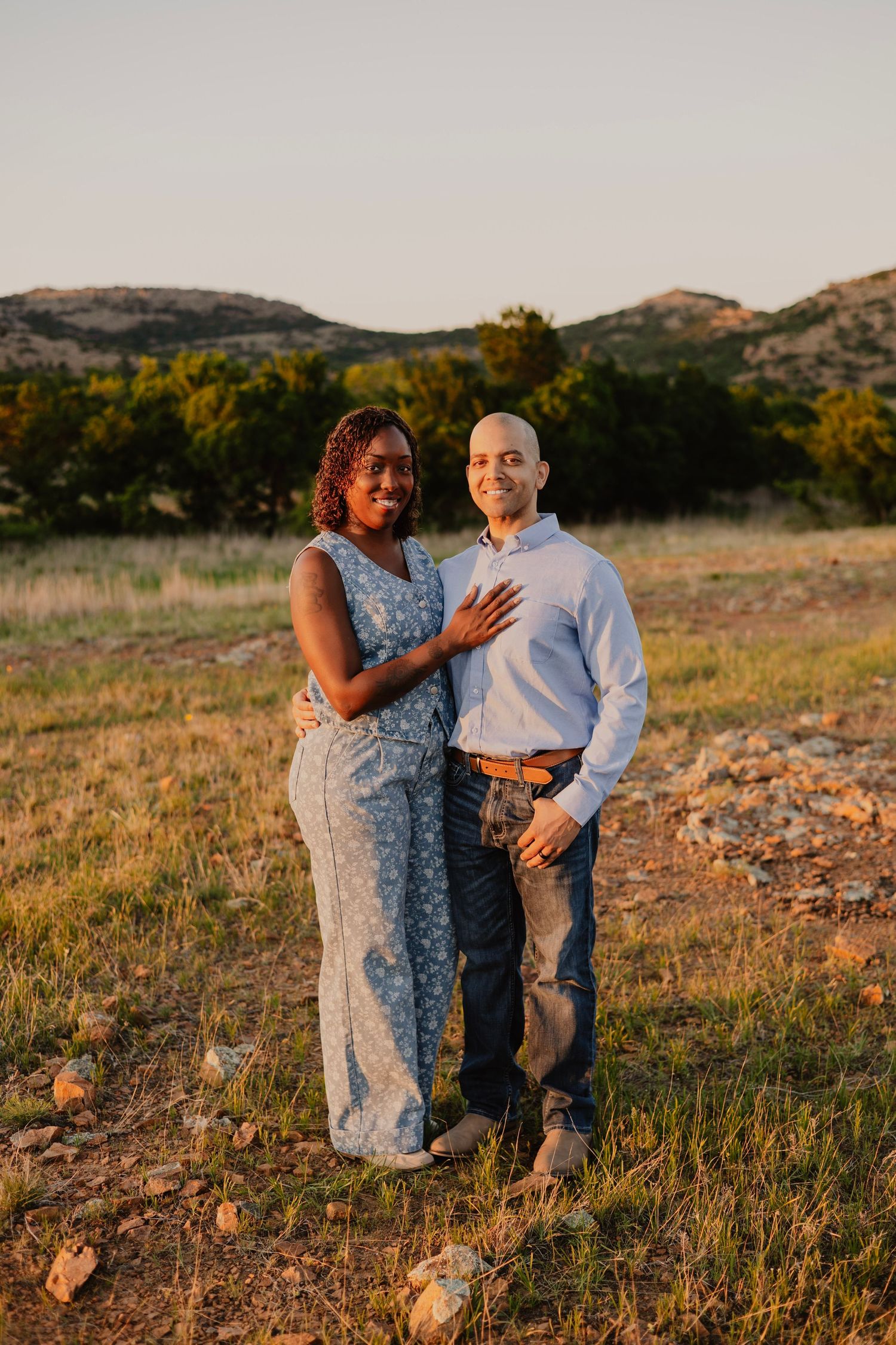 Couple posing at golden hour in a scenic field with hills and trees, woman in floral dress, man in light blue shirt.
