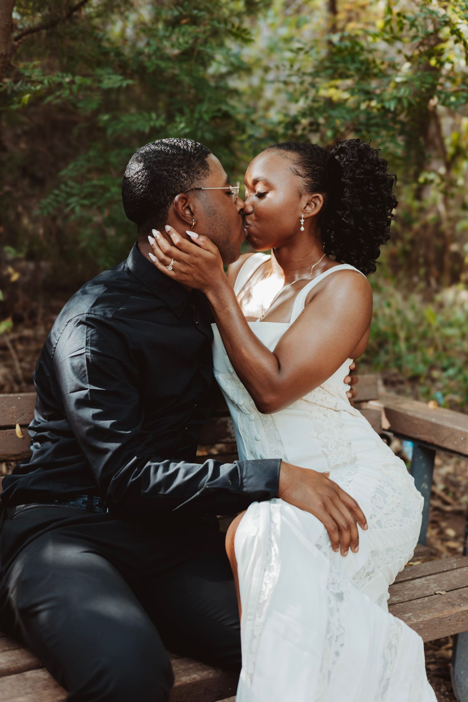 Newlyweds sharing a tender kiss while sitting on a wooden bench in a garden setting.