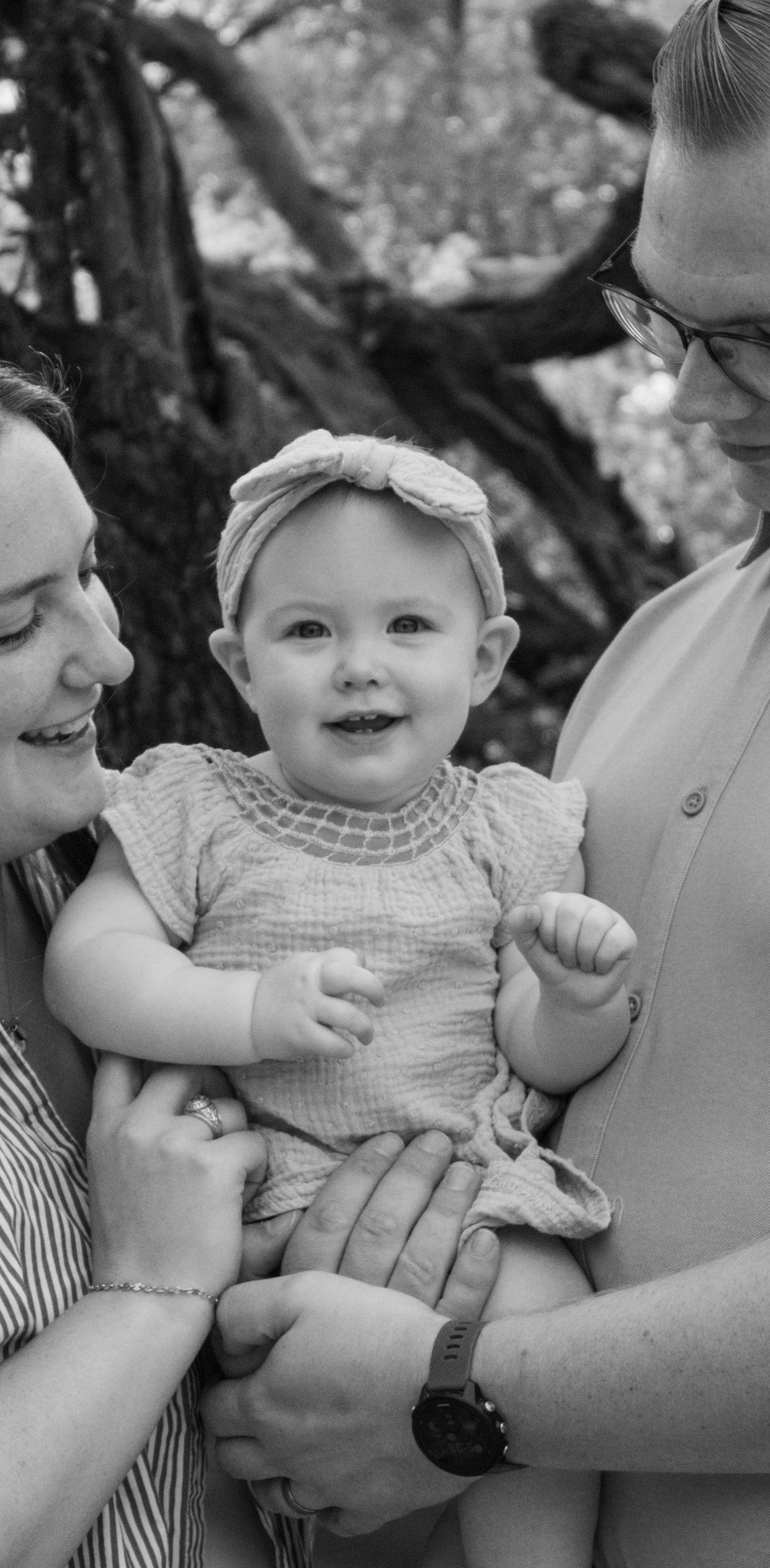 Black and white portrait of parents holding smiling baby with headband.