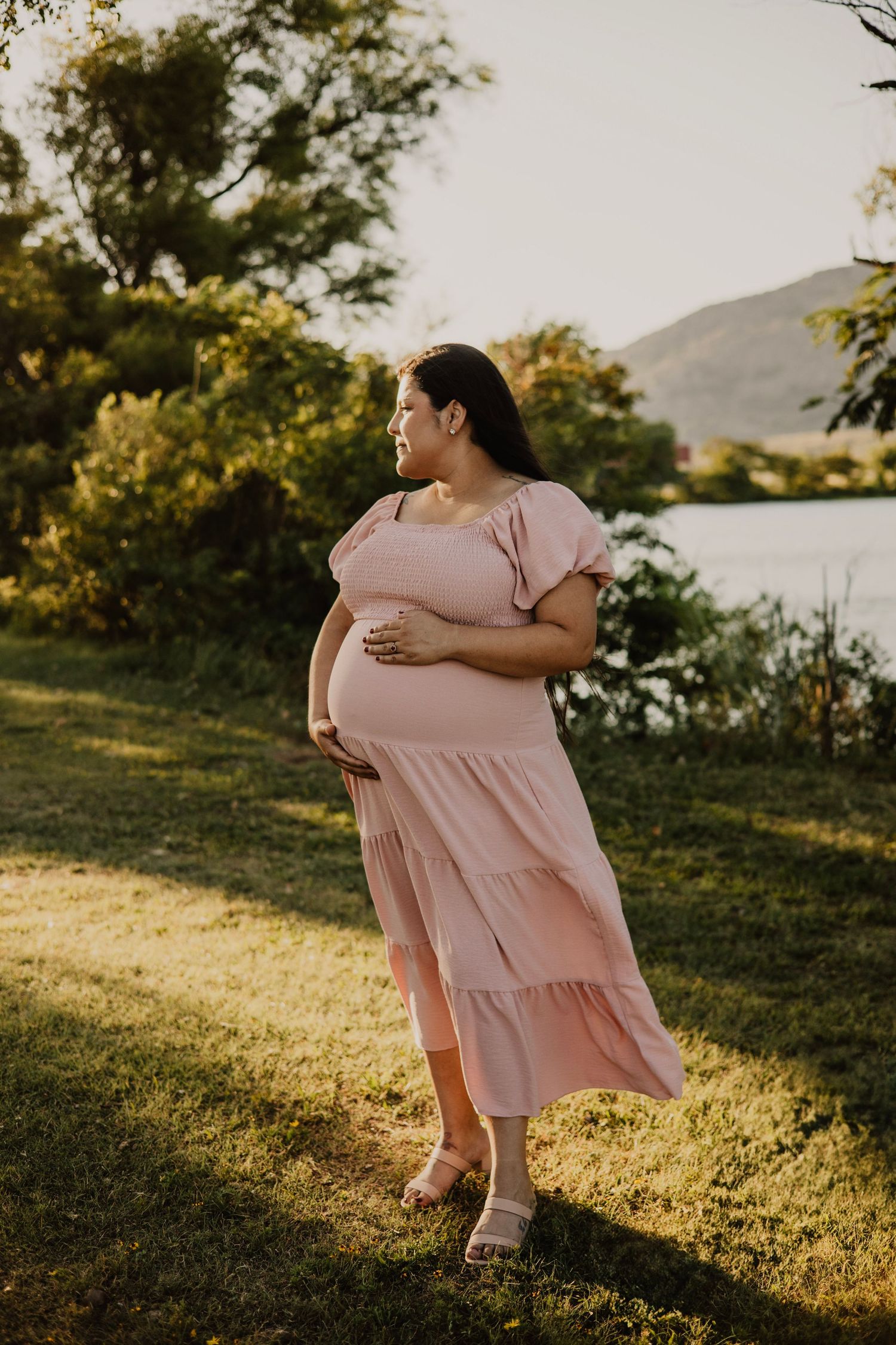 An expectant mother in a flowing pink dress stands gracefully in golden afternoon light by a lake.