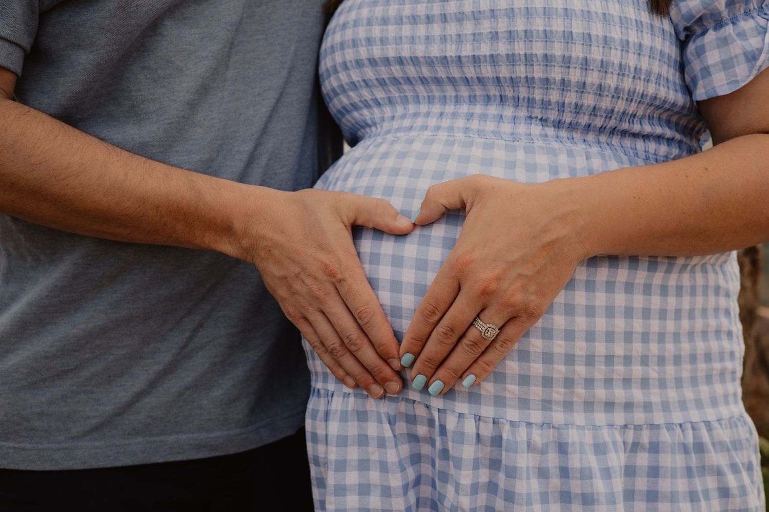Hands forming a heart shape over a pregnant belly in a blue gingham dress.