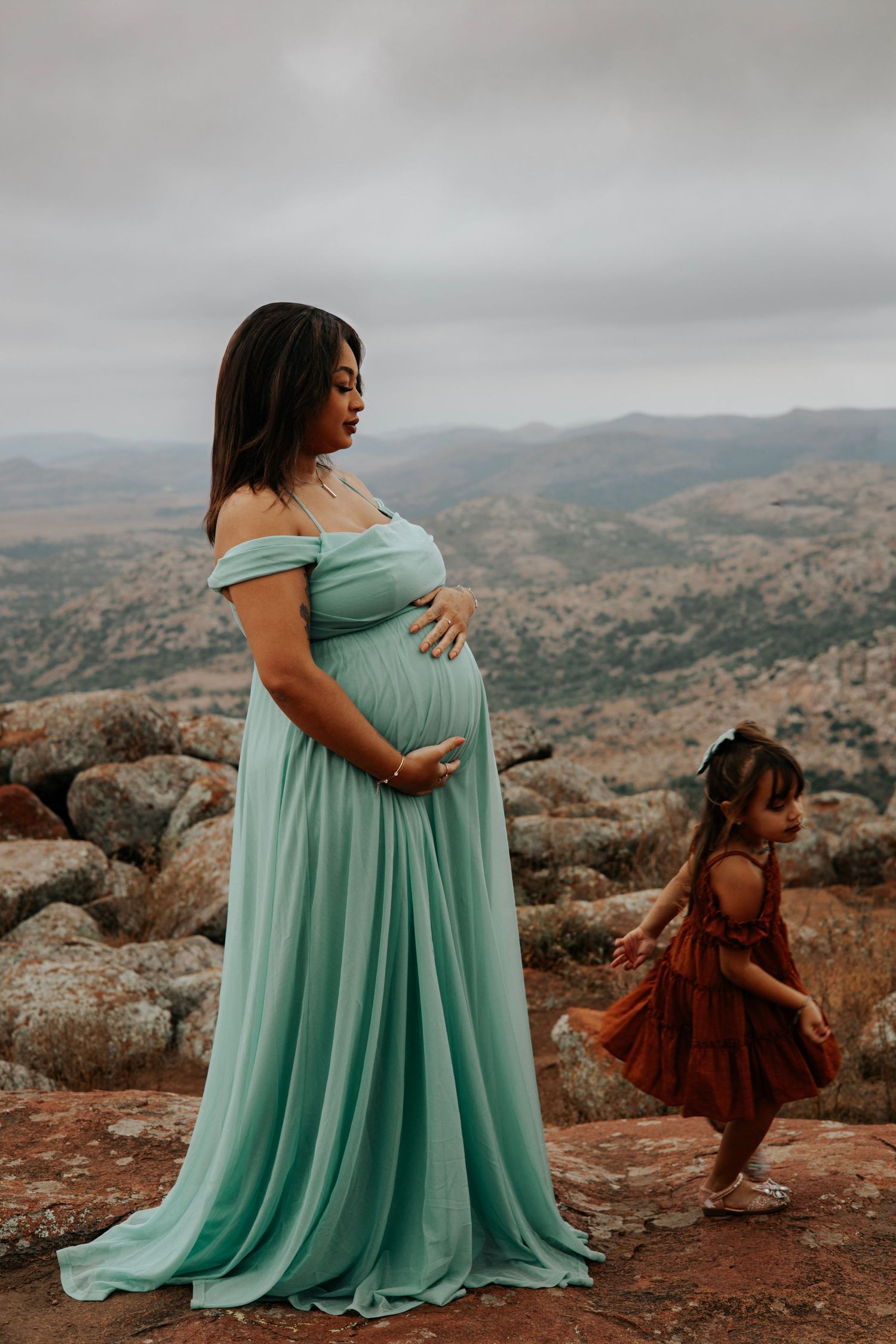 A pregnant woman in flowing mint dress stands on rocky overlook while child plays in background.