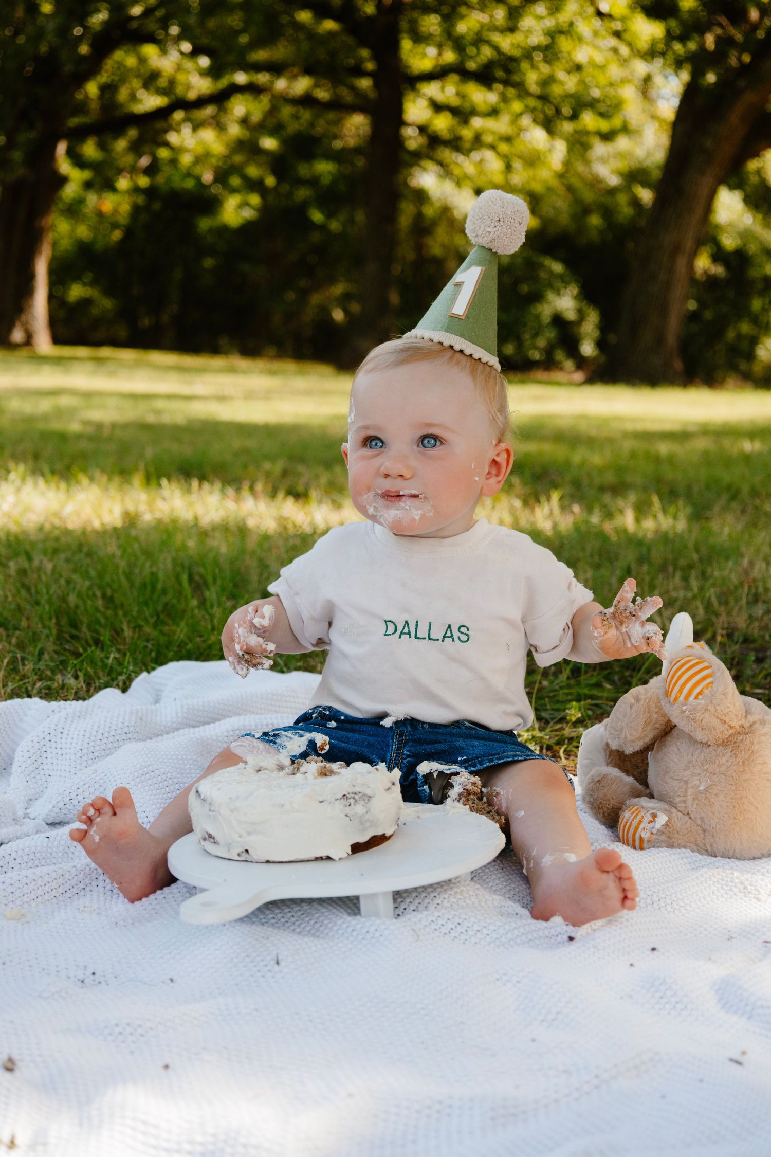 Baby in Dallas shirt and party hat sits on blanket with white birthday cake outdoors.
