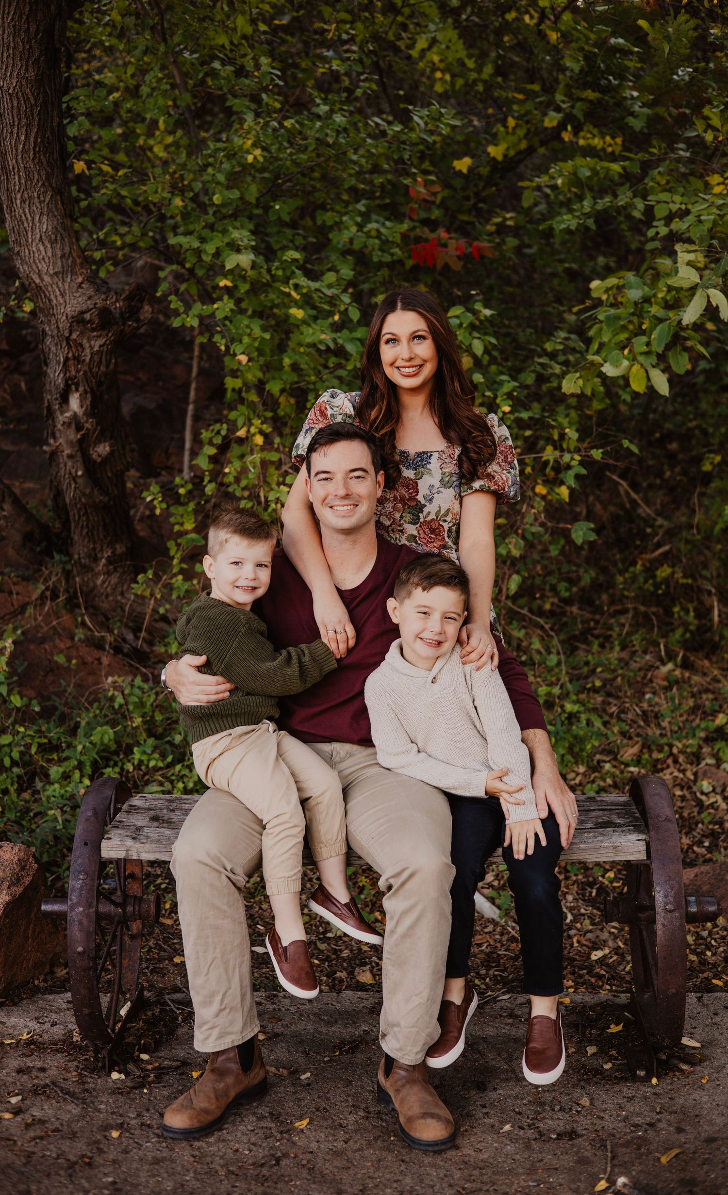 Family poses together on wooden bench with green foliage background.