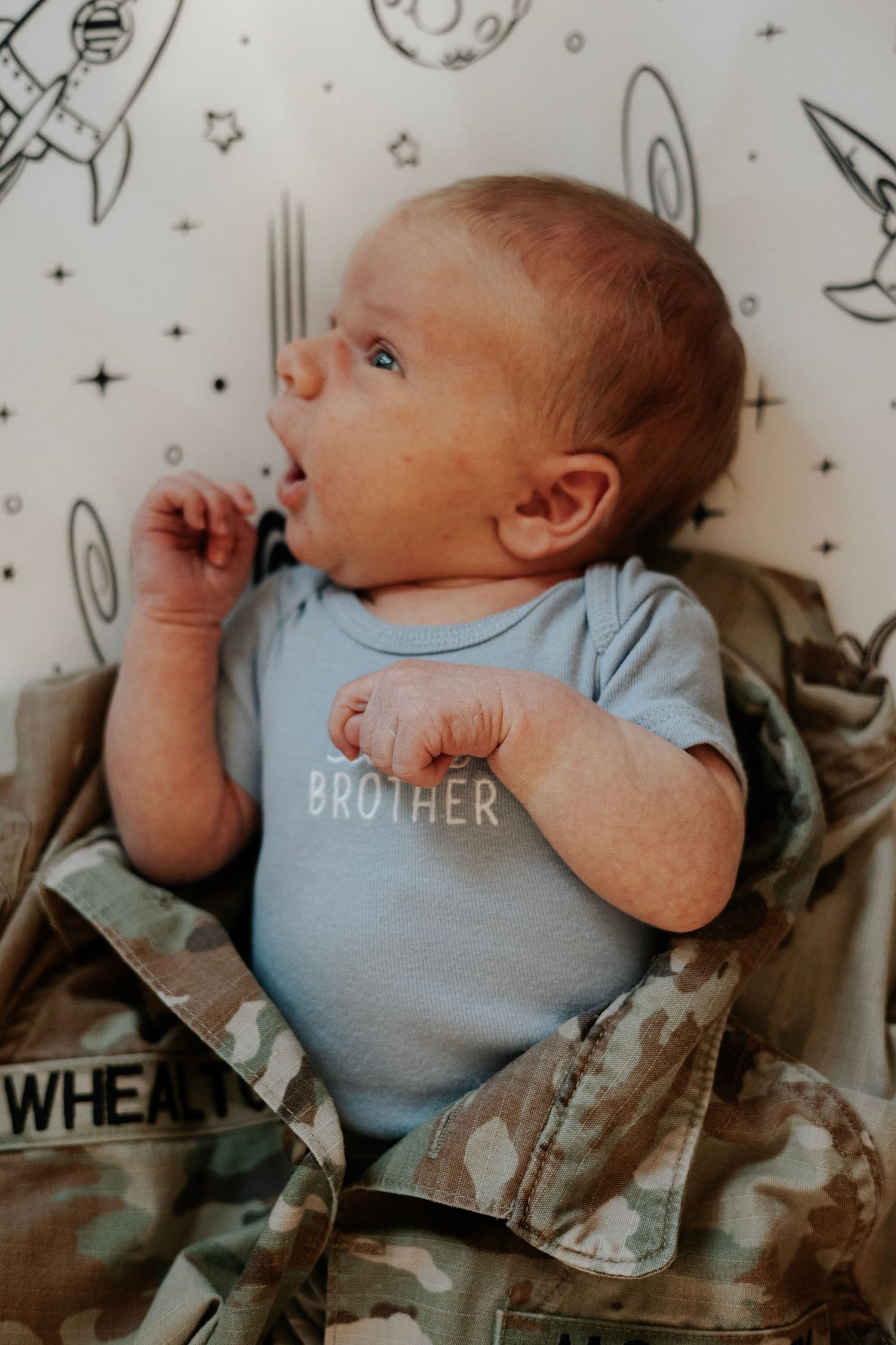 Baby wearing light blue onesie lying on space-themed patterned background.