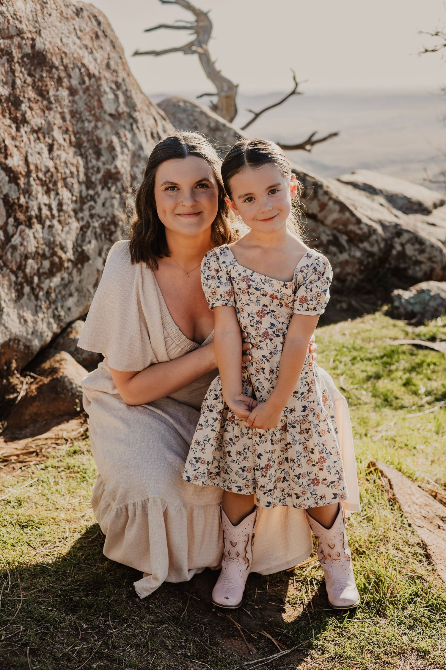 A woman and young girl in floral dress and pink boots pose together on rocky terrain outdoors.