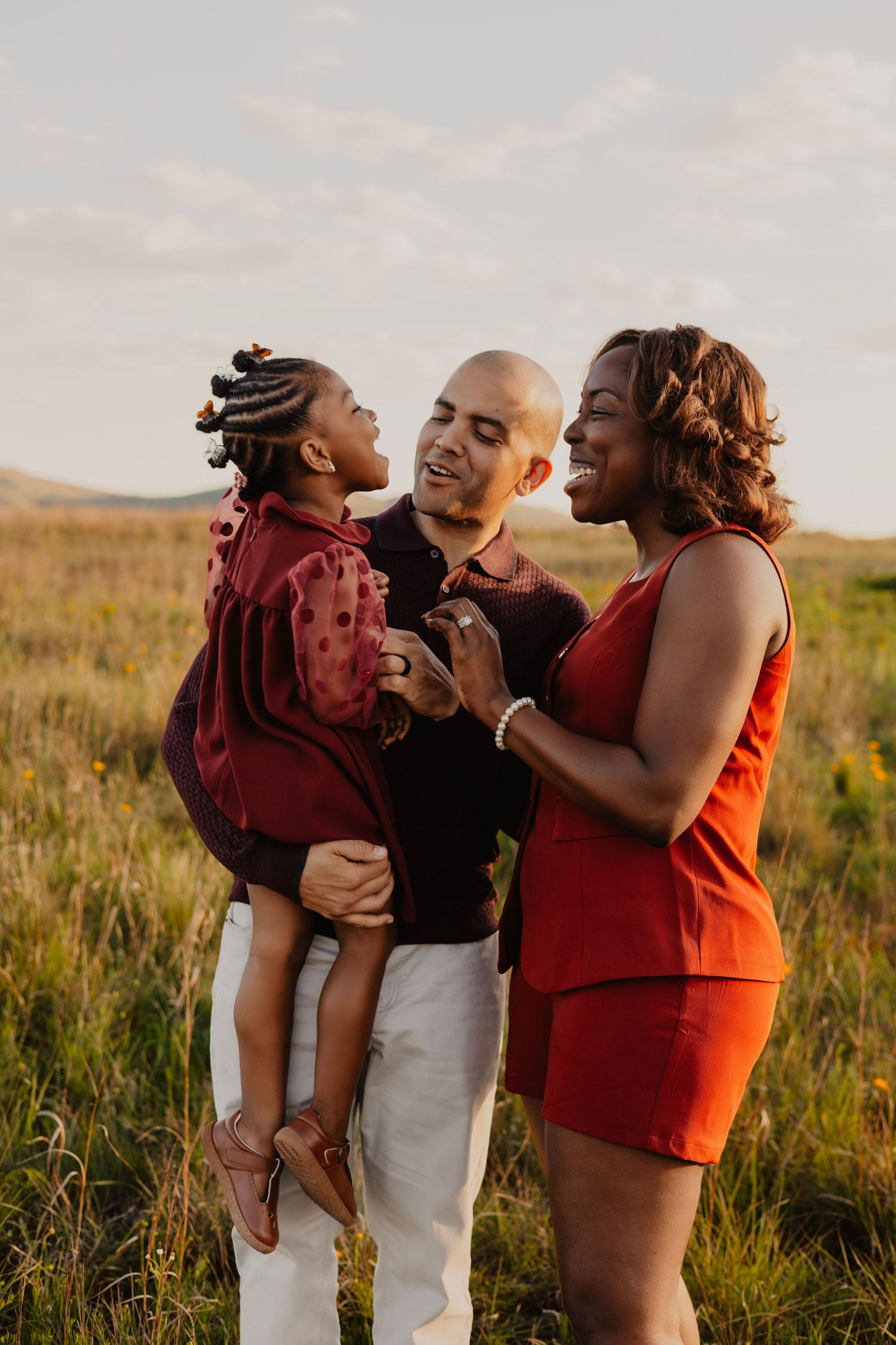 A joyful family of three laughs together in a golden field at sunset, dressed in red and dark tones.