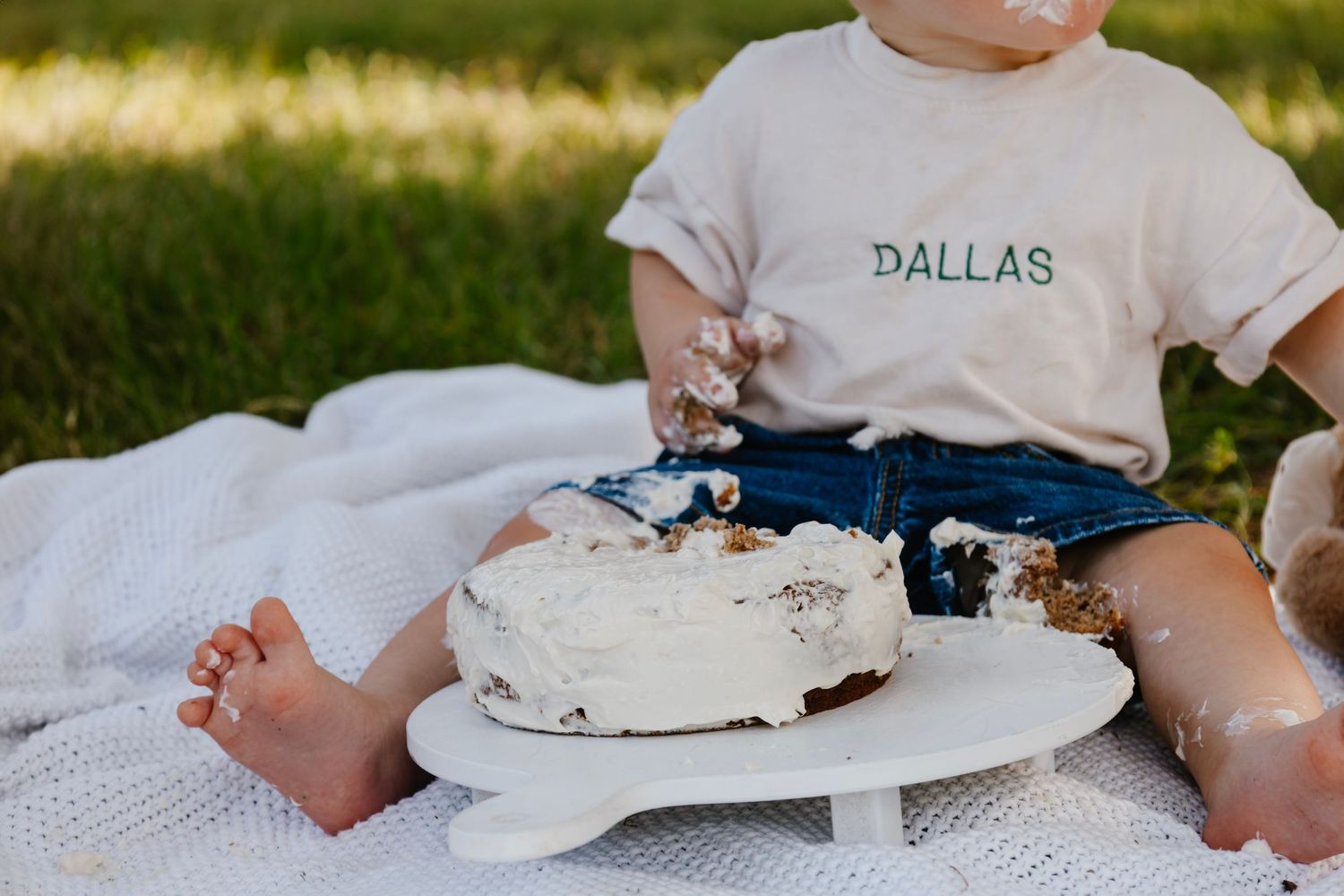 Small child wearing Dallas shirt sits on white blanket with smashed cake during outdoor photo session.