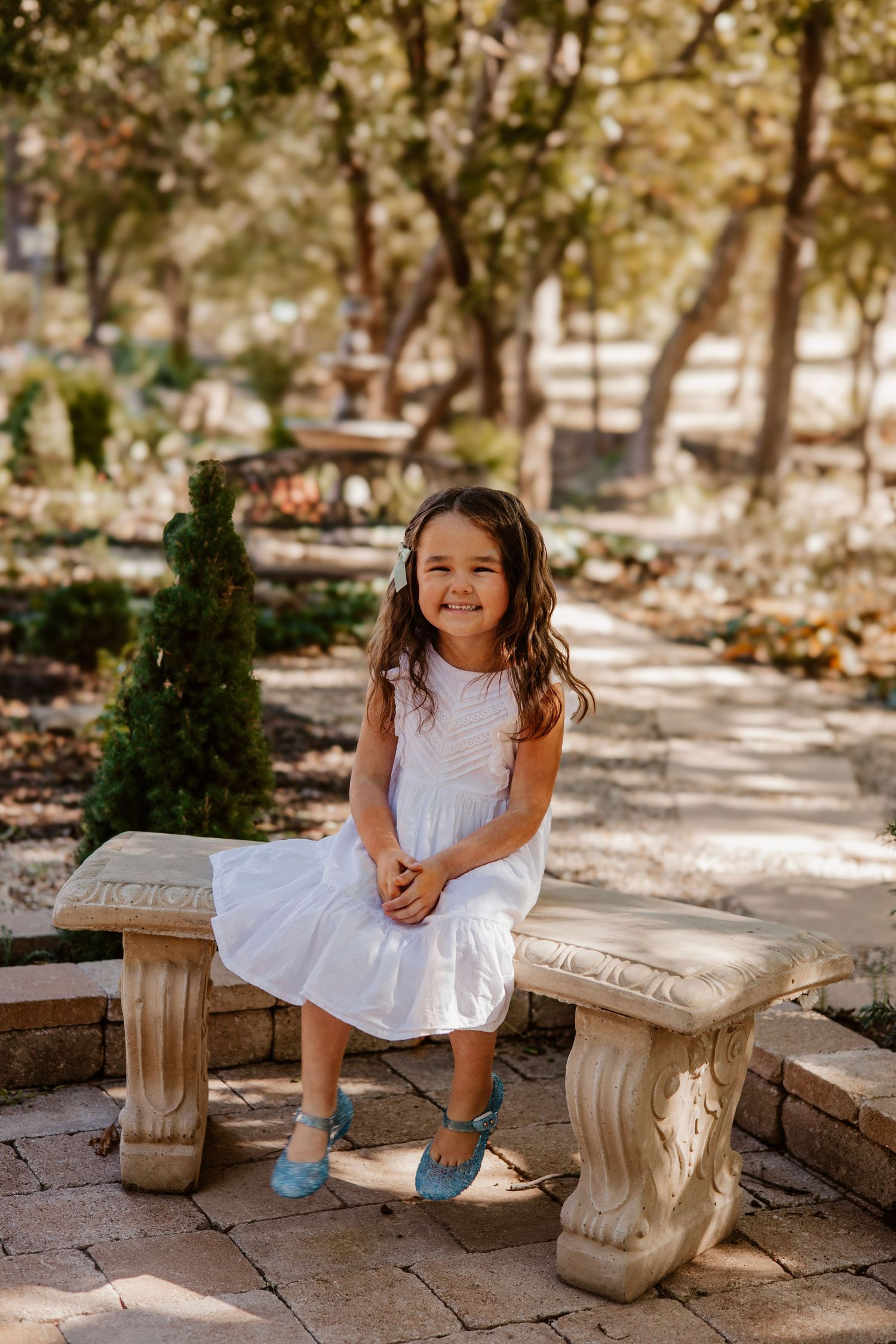 Young girl in white dress sitting on stone bench in garden setting.