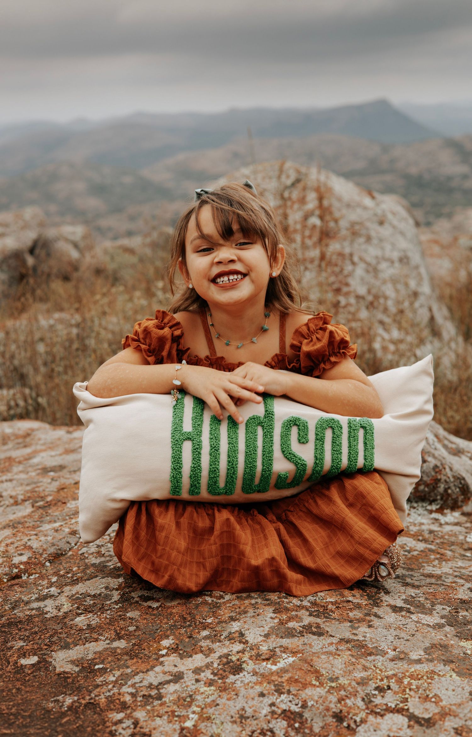A child sits on rocky terrain holding a decorative pillow with 'Hudson' text.