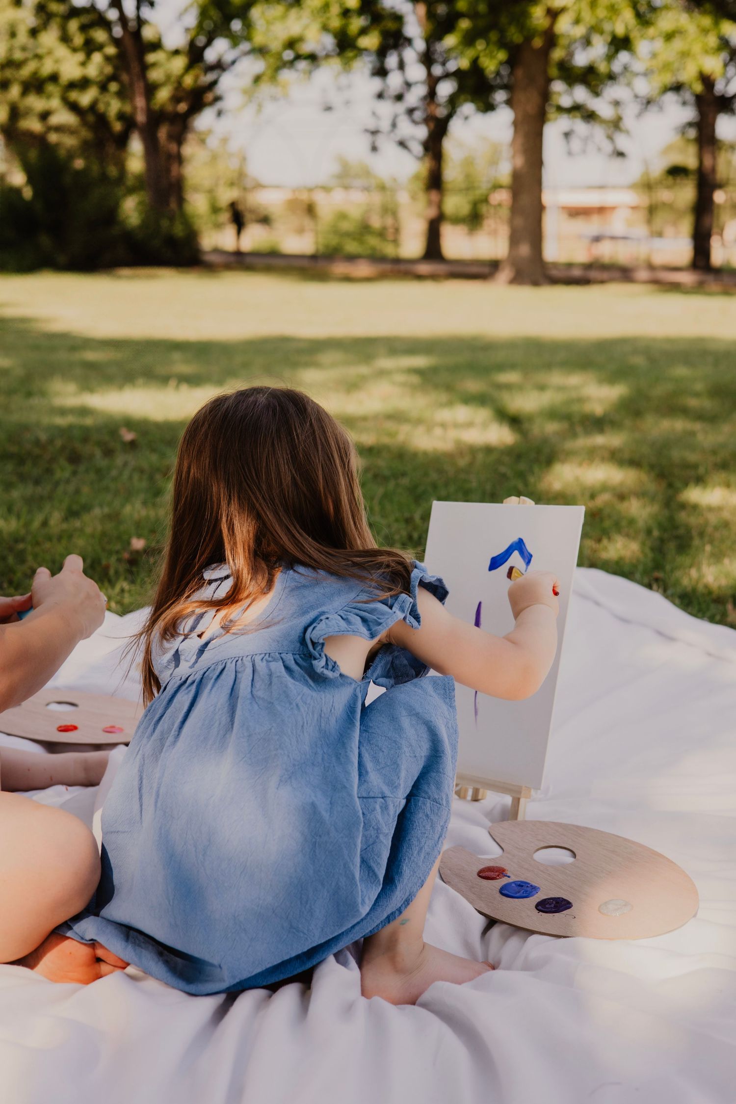 A person in a blue top lies on a white blanket in a sunny park setting while painting or drawing on paper.