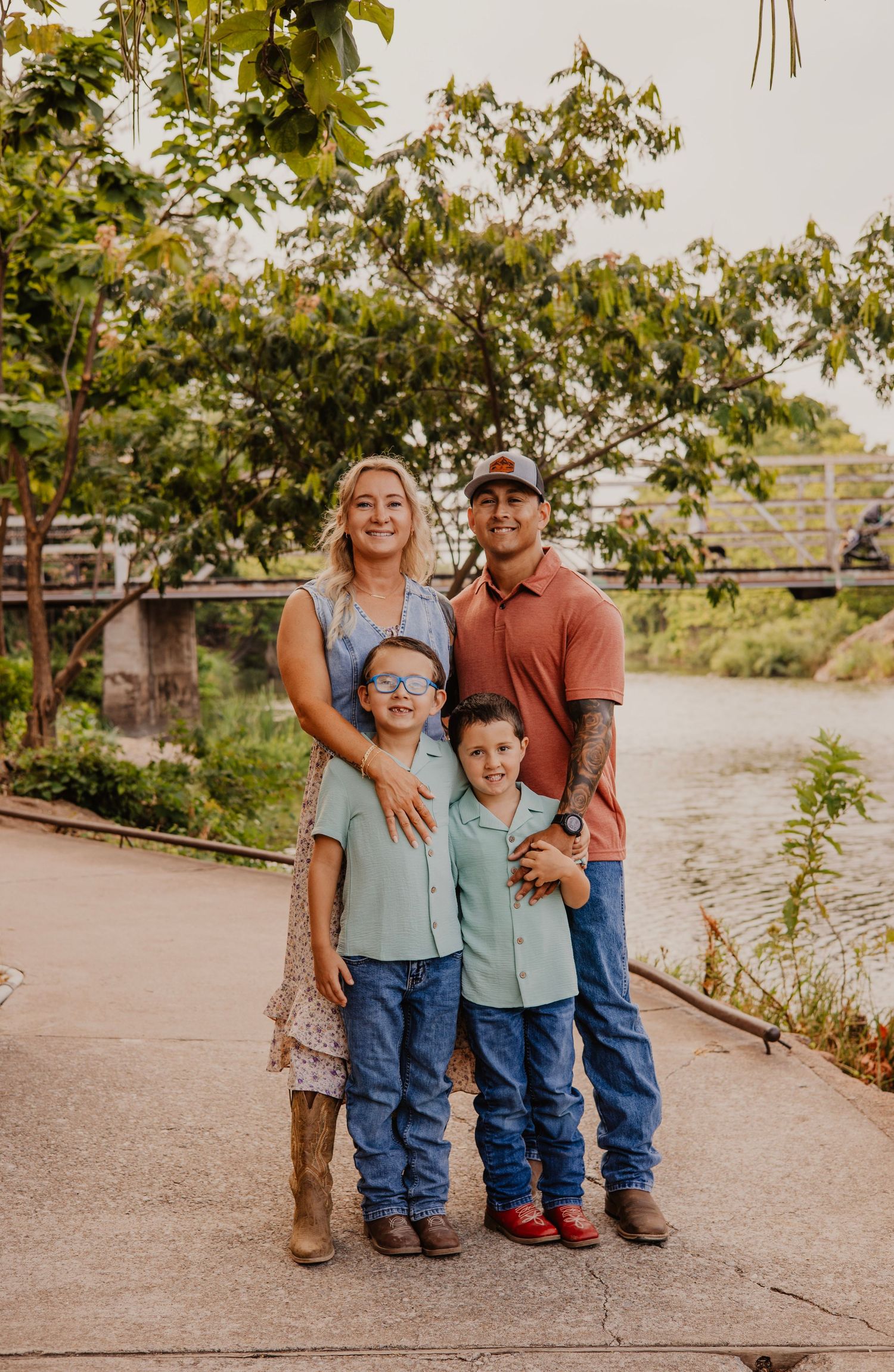 A family of four poses together on a pathway near water with trees in the background.