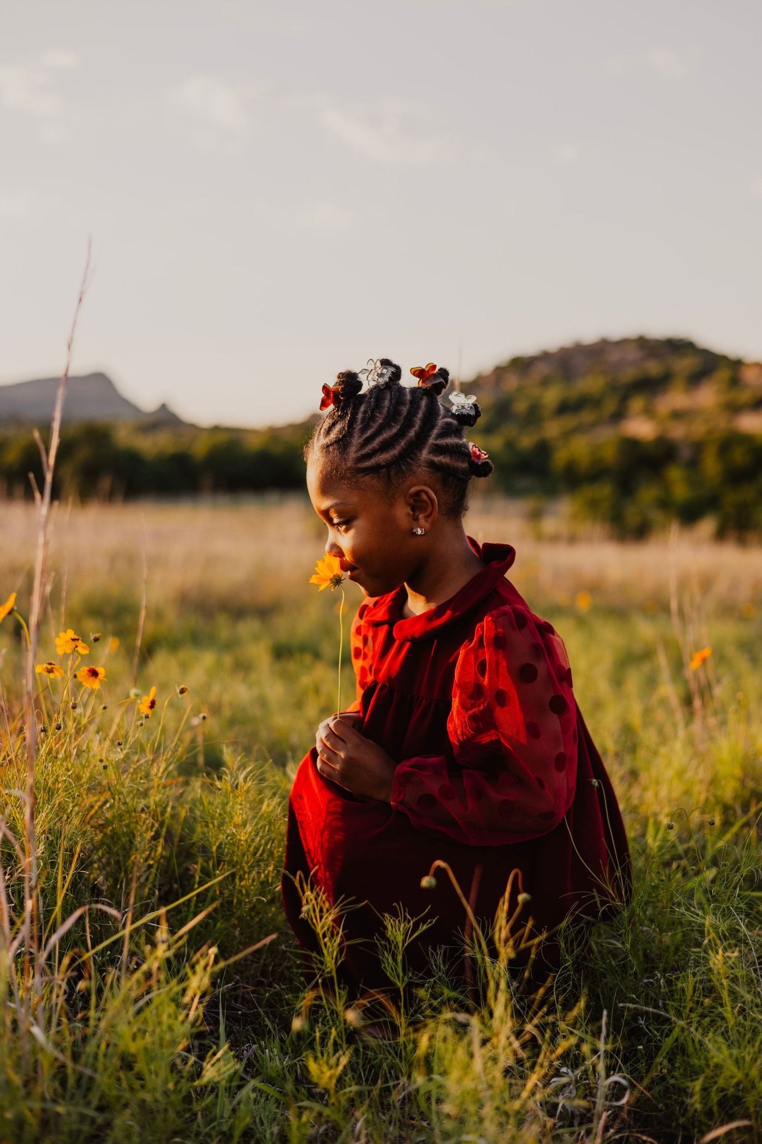 A young Black girl in a red polka dot outfit sits in a golden meadow with wildflowers and hills at sunset.
