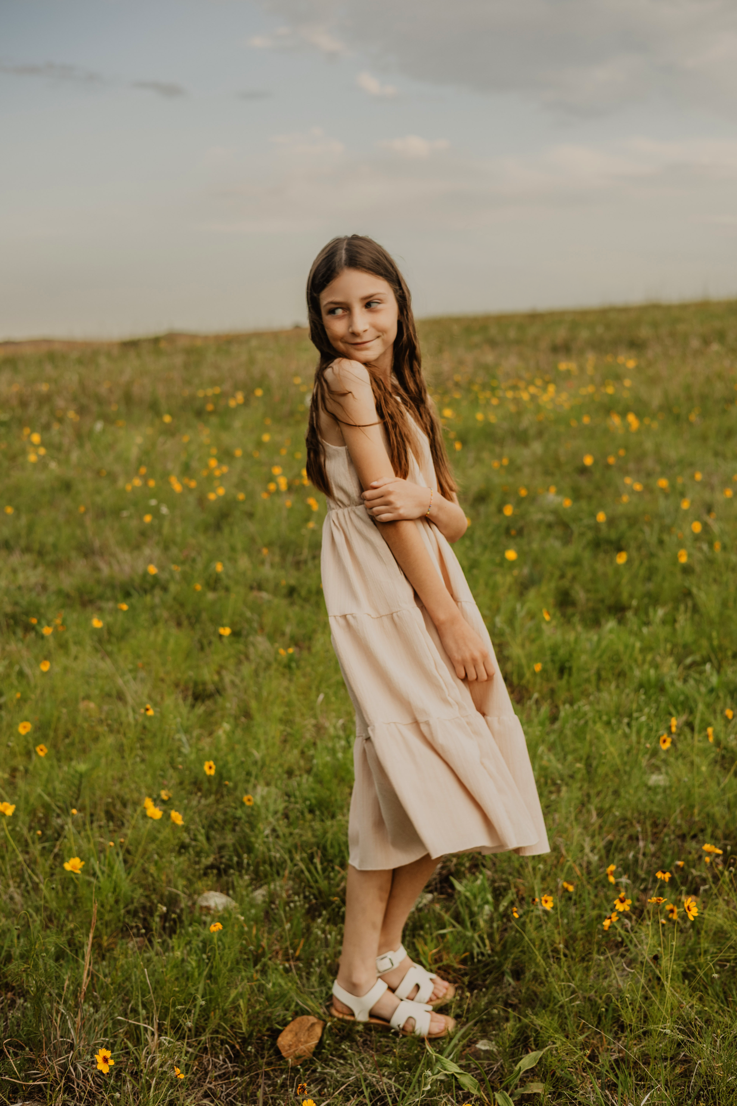 Young girl in beige dress and sandals smiling in a green meadow with yellow wildflowers under cloudy sky.