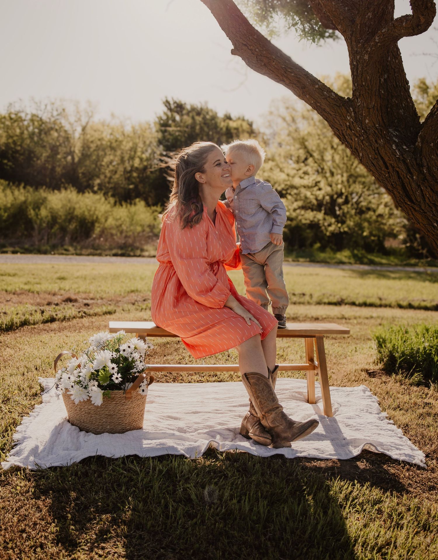 A tender outdoor moment captured at sunset with a coral dress and baby on a rustic bench with flowers nearby.